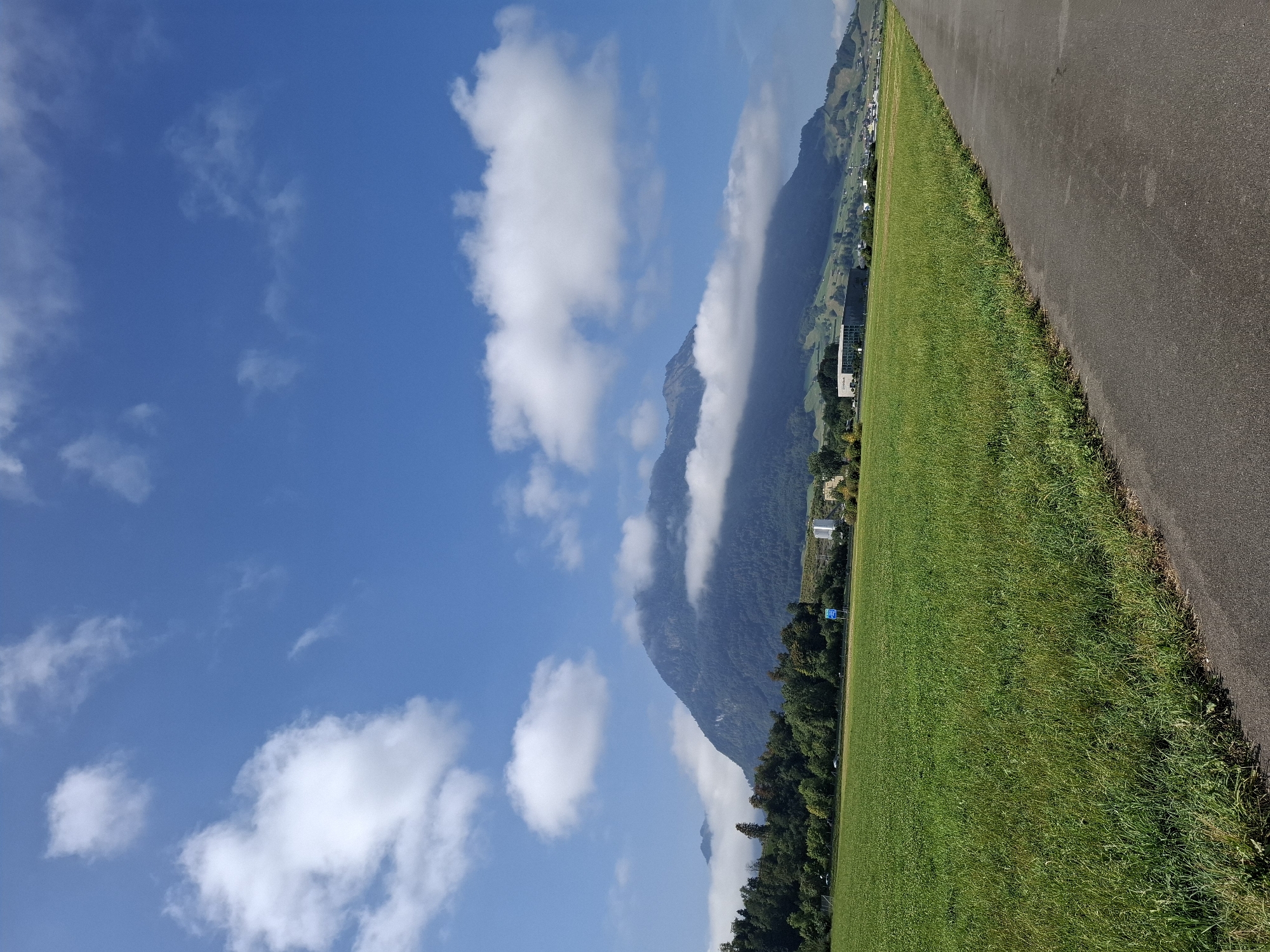 The image depicts a scenic view of the Swiss Alps from the outskirts of Buochs, Switzerland. In the foreground, a paved path runs diagonally from the bottom right corner towards the center of the frame. To the left of the path is a lush, green field of grass that extends into the mid-ground. Beyond the field, a line of trees forms a transition to the mountainous terrain.
The main subject of the image is a large, imposing mountain range that dominates the background. The lower slopes of the mountains are covered in dense forest, while the upper peaks are partially obscured by fluffy white clouds. The sky above is a vibrant blue, dotted with more scattered clouds, suggesting a clear and sunny day.
In the mid-ground, several structures are visible. A modern, multi-story building with a dark facade stands out amongst the greenery on the hillside. Further back, hints of a small town or village can be seen nestled in the valley. There are no visible people or animals in the scene. The overall impression is one of natural beauty and tranquility, with a blend of rural landscape and distant urban development.