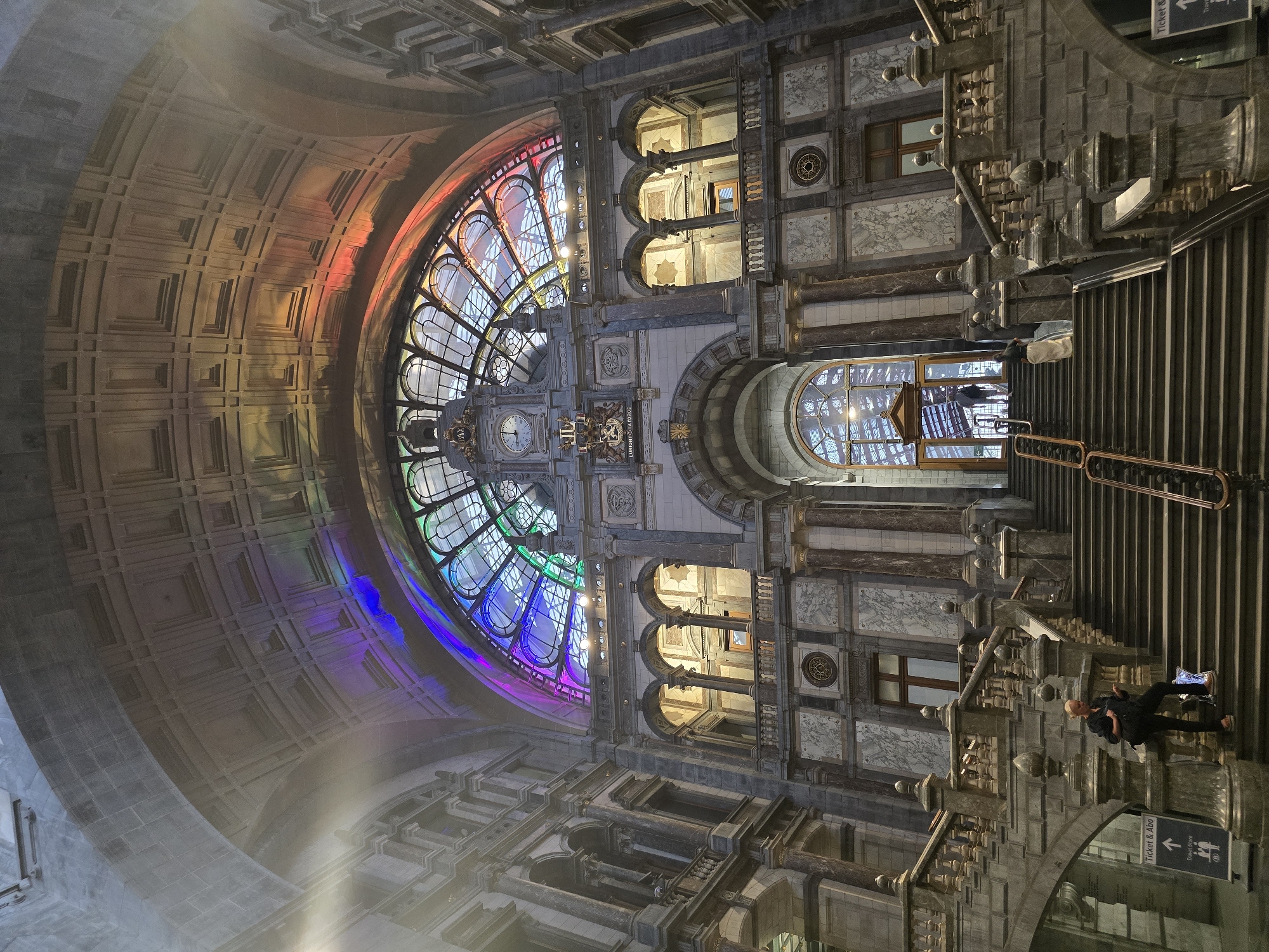 The image depicts the grand interior of the Antwerp Central Station, also known as the "Railway Cathedral," in Antwerp, Belgium. The architectural marvel features a soaring, vaulted ceiling with intricate stonework, illuminated by a spectacular stained-glass window that casts a spectrum of rainbow colors across the space.
In the foreground, a wide, ornate stone staircase ascends, flanked by detailed balustrades and pillars. On the left side of the staircase, a woman stands, looking at her phone, with a bag at her feet. Further up the stairs, two individuals are visible walking towards a large, arched glass entryway.
The station's interior showcases classical architectural elements, with marble accents, arched windows, and grand columns. The overall ambiance is one of opulence and grandeur, enhanced by the dramatic play of light and color from the stained-glass ceiling. The scene is captured from a low angle, emphasizing the scale and magnificence of the station.