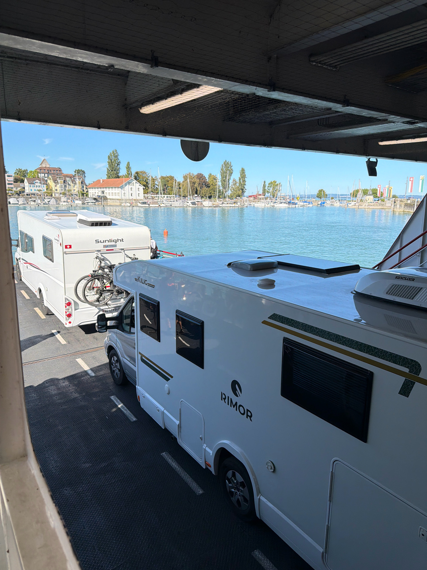 The photograph captures a scene on a ferry crossing Lake Constance, with the city of Romanshorn, Switzerland, visible in the background. In the foreground, two white recreational vehicles (RVs) are parked side-by-side on the ferry deck. The RV closest to the viewer is a Rimor model, and its front half is visible, showing its roof and part of its side. The second RV, further back, is a Sunlight model, and its entire rear section is visible, including a bicycle rack mounted on the back with a bicycle attached.
The ferry deck is a dark, textured surface, marked with white lines and reflective diamond-shaped indicators. Above the vehicles, the underside of the ferry's roof is visible, featuring industrial lighting and netting.
The background offers a picturesque view of Romanshorn. Buildings with red-tiled roofs and yellow and white facades line the waterfront. A marina with numerous sailboats is prominent, with masts extending into the bright blue sky. Several flags are visible on poles along the shore, suggesting a public or official area. The water of Lake Constance is calm and a vibrant blue. The overall lighting suggests a bright, sunny day. There is no specific indication of any people interacting with each other or the vehicles, other than the presence of a figure in the distance near the ferry railing.