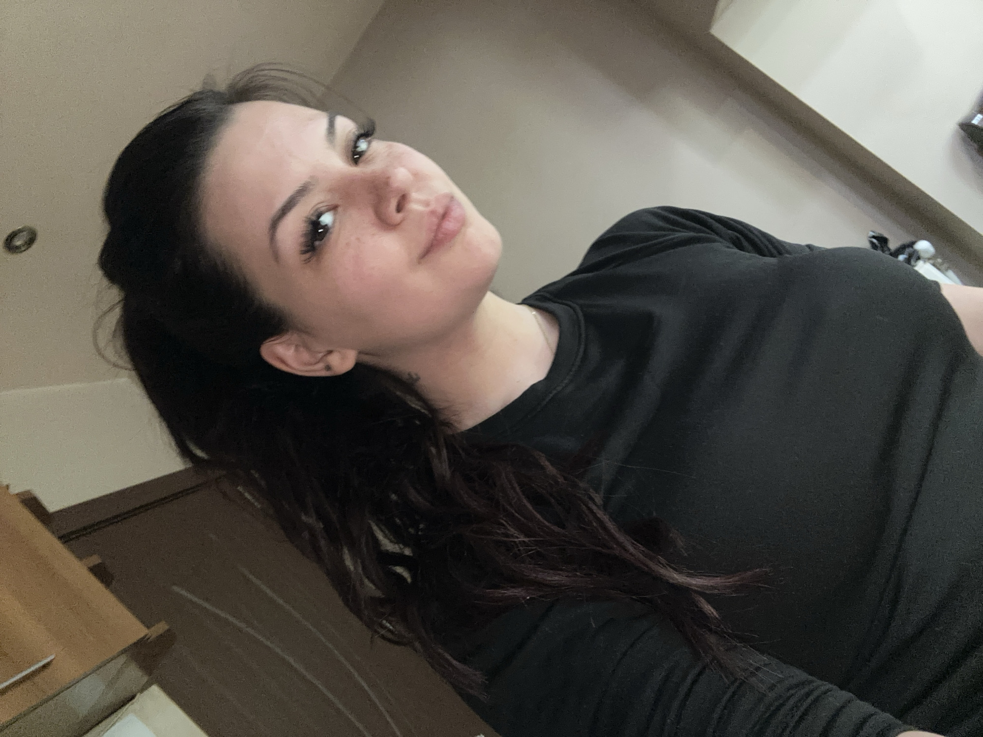 A young woman with dark hair pulled back in a ponytail poses for a selfie. She is wearing a long-sleeved black shirt. Her head is tilted slightly and she is looking at the camera with a slight smile. Her background includes part of a wooden door, a light-colored wall and ceiling. The image does not provide sufficient information to determine its location within Sliven, Bulgaria.