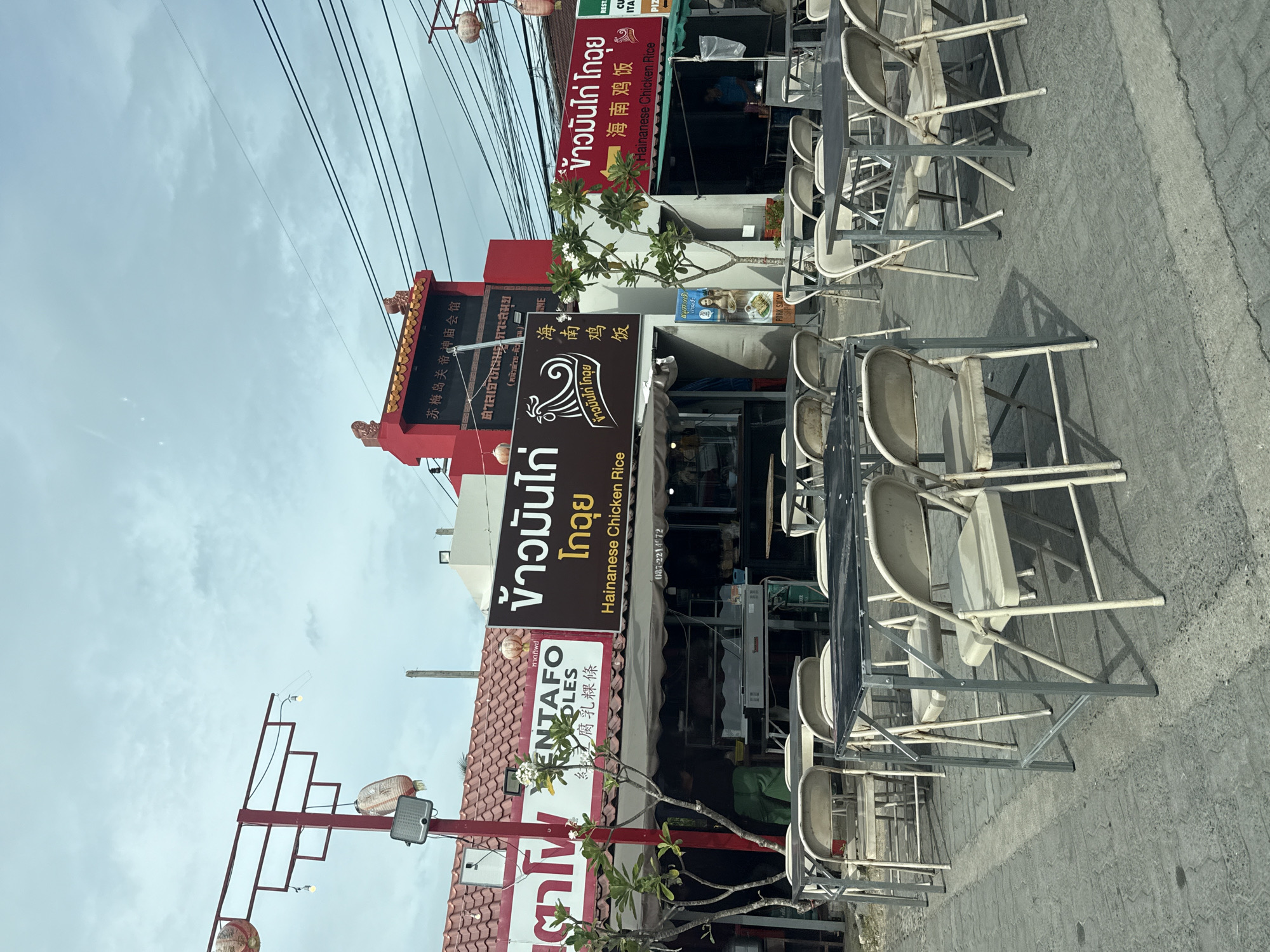 This is a daytime, eye-level, medium shot of an outdoor dining area in front of several shops on the island of Ko Samui, Thailand. The foreground is dominated by metal tables and simple, folding chairs set up on a paved surface. Behind the tables, storefronts display signage in Thai and Chinese. One sign prominently advertises "Hainanese Chicken Rice." The buildings are modest, with red tile roofs and awnings. A young woman in green pants stands in the doorway of one shop, and another person, presumably an employee, is visible further inside. A leafy green tree stands between two of the shops, and overhead, power lines crisscross the sky, which is overcast. Chinese lanterns hang from red poles, adding a decorative touch to the streetscape. The overall impression is of a casual, local eatery in a tropical setting.