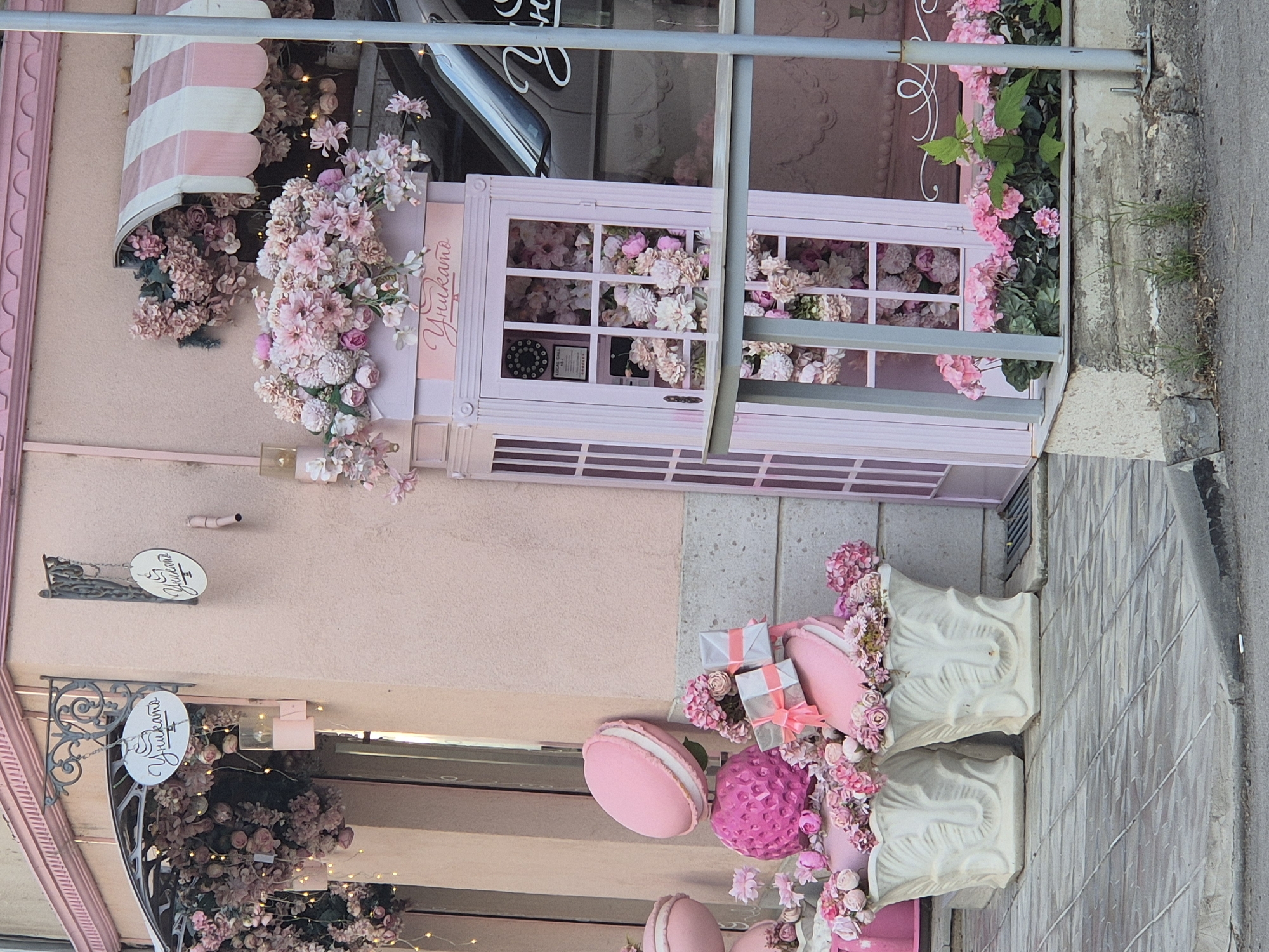 The image shows the facade of a shop in an unknown city. The shop is decorated in pink and white. There is a pink awning over the window and a sign with the name "Yunakamo" on it. There are also flowers and other decorations in front of the shop.