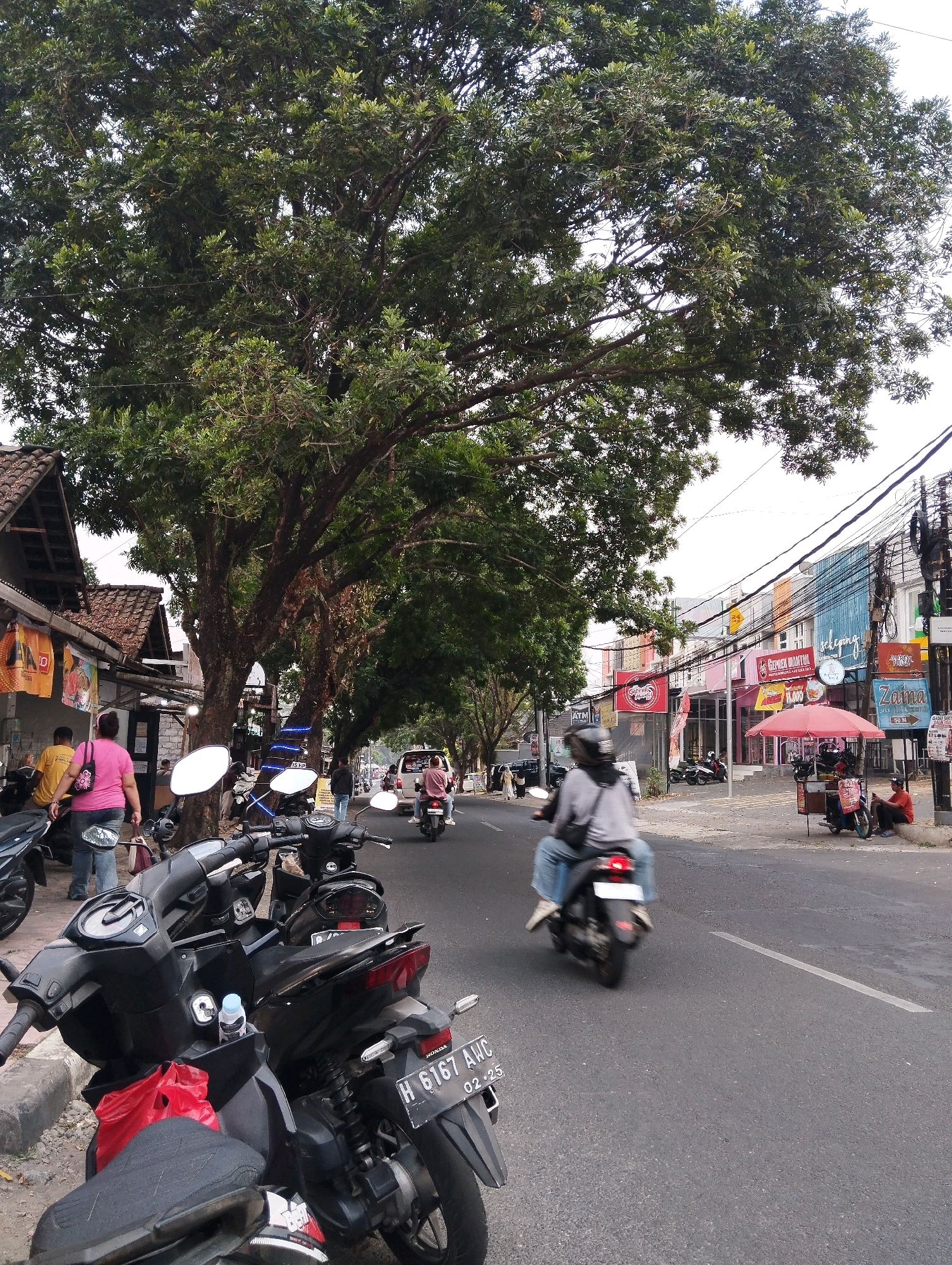 The image depicts a street scene in Salatiga, Indonesia, characterized by a narrow road lined with businesses and large trees. In the foreground, several parked motorcycles are visible, one with a red plastic bag hanging from its handle. A woman in a pink shirt and jeans is walking away from the viewer towards a building. Further down the street, a motorcyclist is riding away from the camera. On the right side of the road, a red umbrella shades a vendor cart. Various shopfronts with colorful signage, including "Zaina," are visible along the street. Electrical wires are strung across the scene. The overall atmosphere suggests a busy urban environment with a mixture of commercial activity and daily life.