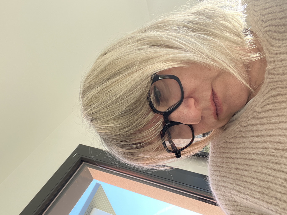 The image displays an adult female, likely indoors within Chambœuf, France. She is seen in a close-up, slightly upward-angled perspective. The woman possesses shoulder-length, straight blonde hair with bangs. She is wearing large, black, square-framed glasses, through which her eyes are visible, directed downwards. Her mouth is closed. She is dressed in a light beige, ribbed knit sweater, and a thin silver chain is faintly visible at her neckline. In the upper left background, a dark-framed window reveals a clear blue sky, part of a white building structure, and a reddish-brown exterior wall. The upper right portion of the background consists of a plain, light-colored wall or ceiling. No direct interactions are depicted.