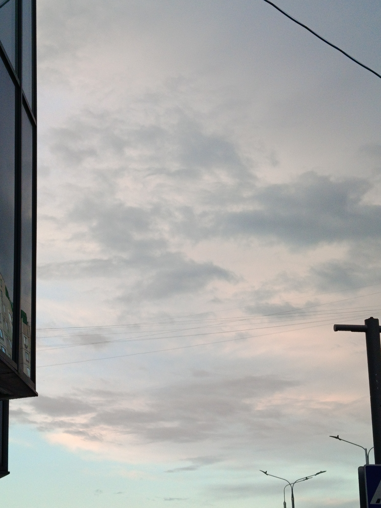 The image depicts a cloudy sky above a city scene. A portion of a modern building with reflective glass windows is visible on the left. Electrical wires stretch across the sky. On the right, there is a dark-colored utility pole and a street light. The sky is filled with layered clouds, showing hues of gray, white, and hints of pink and blue. The environment suggests an urban area, potentially in Moscow, Russia, given the style of architecture and street infrastructure. There are no people visible in the shot.