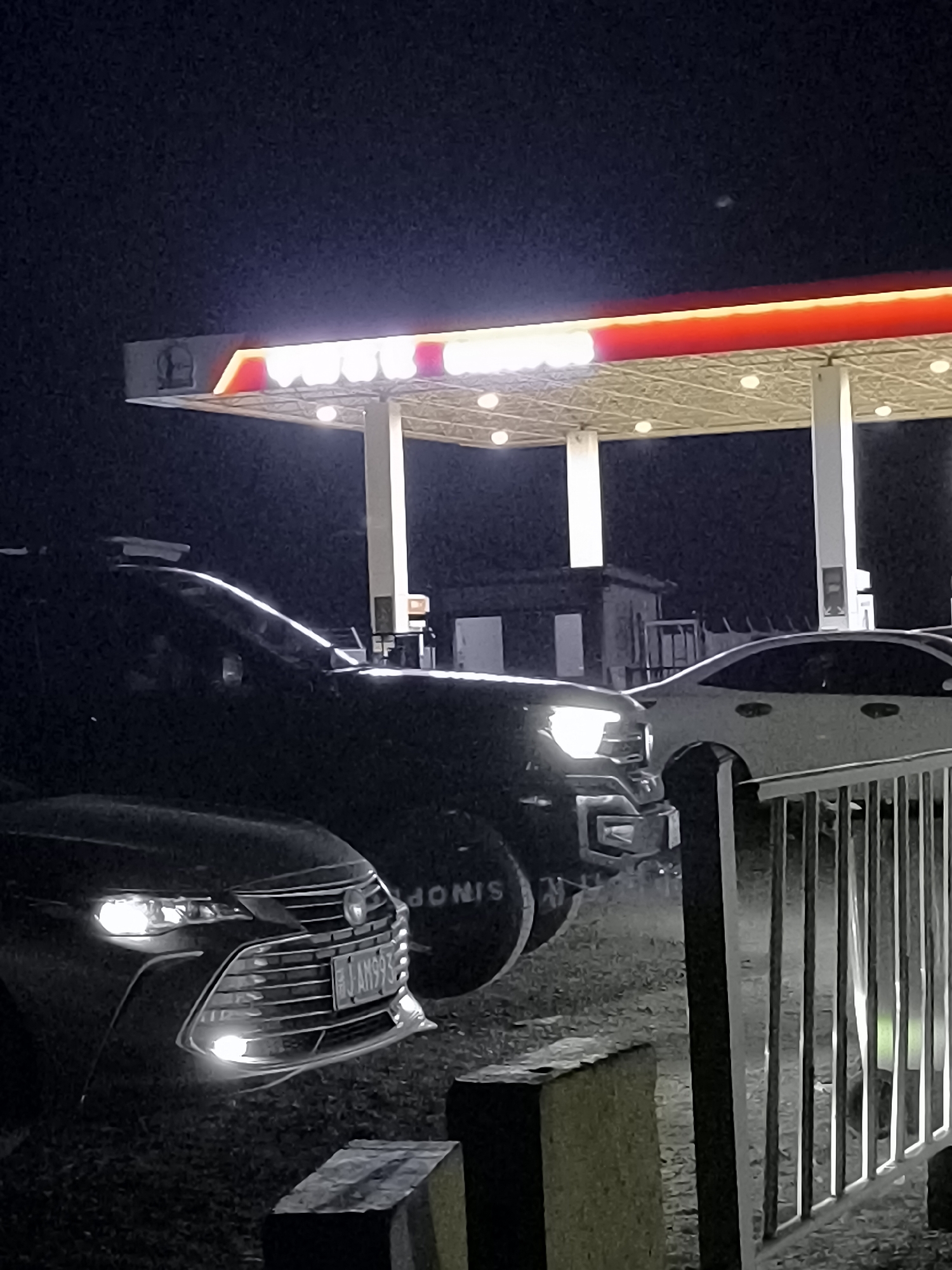 At night, at a gas station in Gurtu Town, China, several vehicles are parked beneath a brightly lit canopy. A dark SUV is parked to the left of the frame, its headlights on, illuminating the front of a dark sedan parked to its left. The sedan has a license plate that reads "J AM993". In the background, a white sedan is partially visible, parked near a metal fence. The gas station canopy is illuminated with white lights and has a red trim. The sky is dark and appears to be clear.