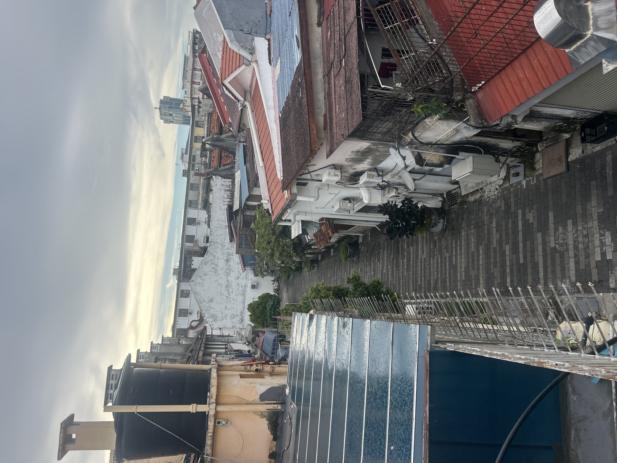 This is an elevated view of a narrow alleyway in George Town, Malaysia. The alley is paved with grey brick pavers and lined with closely packed, two- and three-story buildings. Many of the buildings have red tile roofs, and some feature external air conditioning units and satellite dishes. The architecture appears to be a mix of colonial and traditional Asian styles.
On the left side of the alley, a tall, black water tank sits atop a weathered, yellow-painted building. In the foreground on the left, there is a roof structure with translucent, ribbed panels, possibly a modern addition. The sky is overcast with some hints of yellow and blue, suggesting early morning or late afternoon light. In the distance, a modern, tall skyscraper is visible above the lower buildings. The overall impression is of a densely populated urban environment with a mix of old and new structures. No people are visible in the frame.