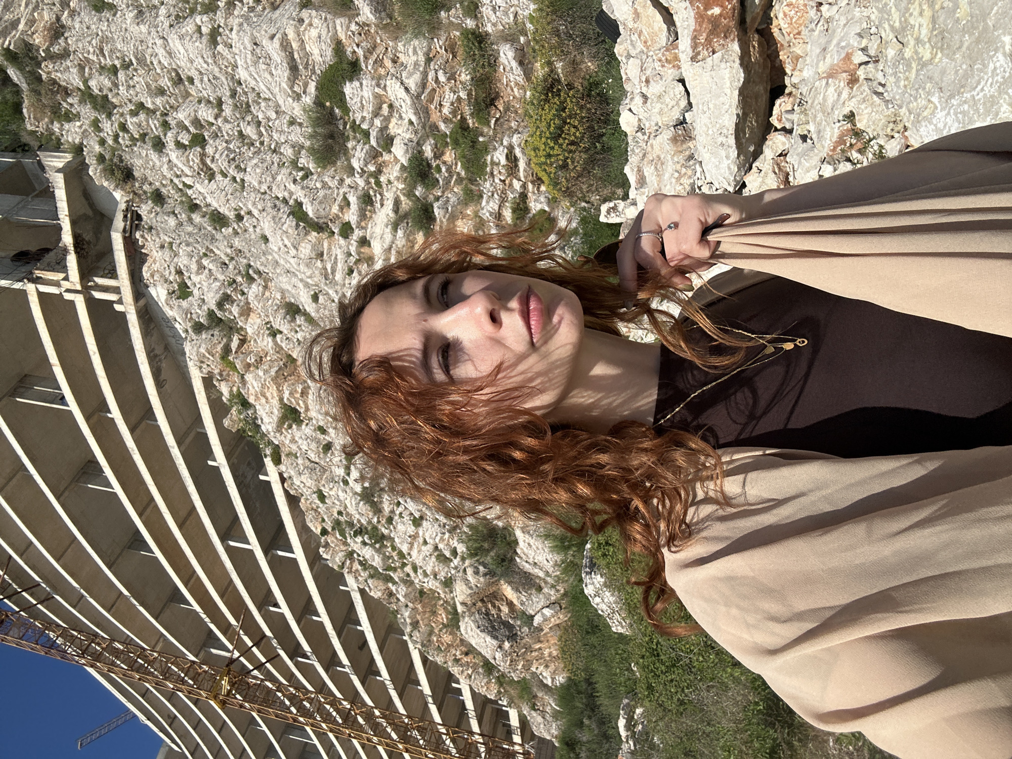 The image shows a woman with red curly hair in Perazića Do, Montenegro. She is wearing a brown shirt and a light beige shawl. She's standing on a cliff with rocks and some sparse green vegetation. In the background, there is a large, unfinished concrete structure, possibly a building or bridge, supported by a crane. The sky is visible and blue in the lower left of the image.
