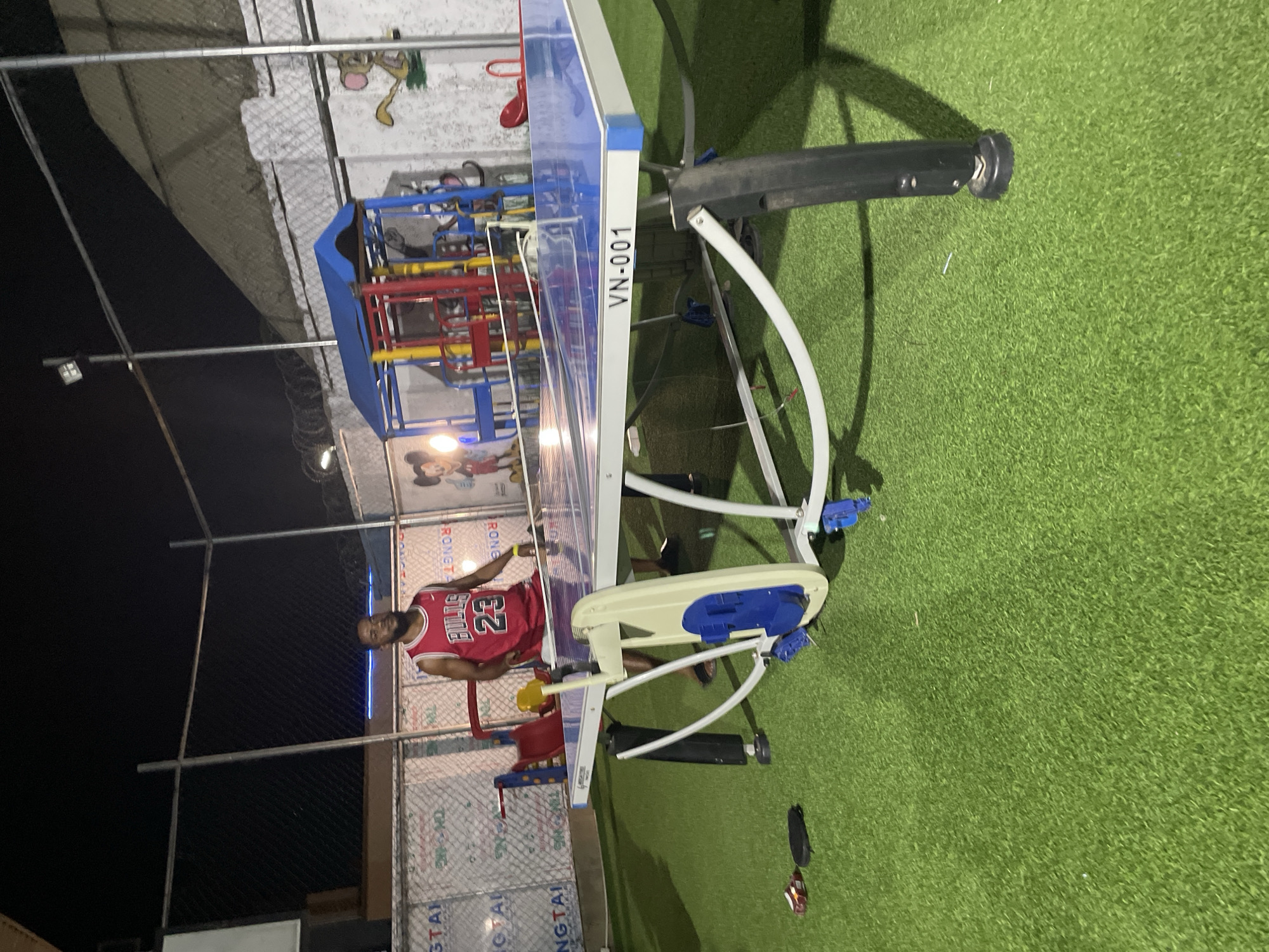 The image depicts an outdoor scene, likely at night, in Akwanga, Nigeria. A man wearing a red Chicago Bulls #23 jersey stands beside a piece of playground equipment. The equipment is painted in bright colors, including blue, red, and yellow. Adjacent to the equipment is a piece of exercise equipment with the marking "VN-001." The ground is covered in artificial green turf. A chain-link fence is visible in the background.