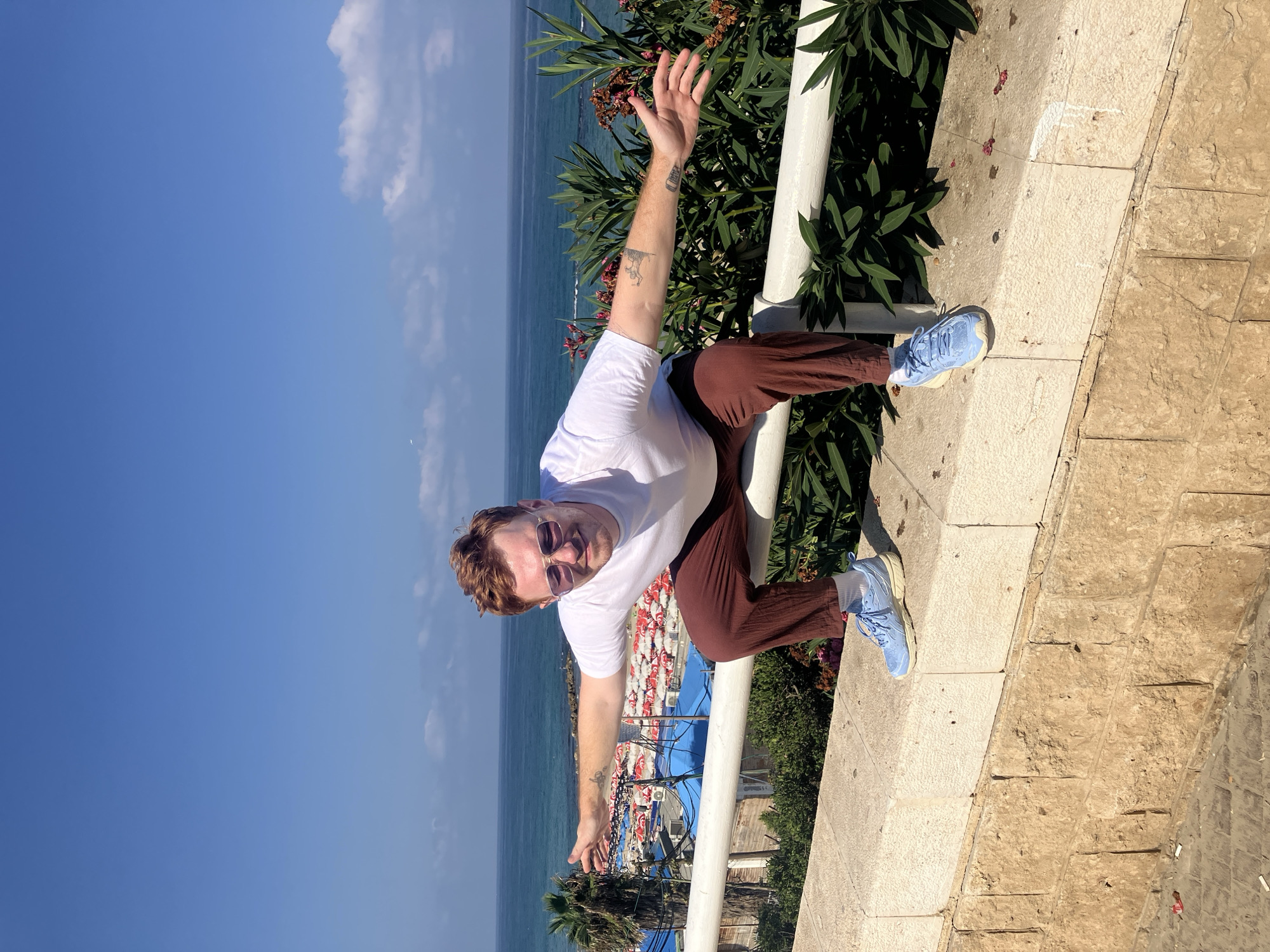 A man with red hair and sunglasses is crouching on a stone wall overlooking the Mediterranean Sea in Bat Yam, Israel. He is wearing a white t-shirt, brown pants, and light blue sneakers, with his arms outstretched to the sides. Behind him, a beach with red and white umbrellas is visible, as well as palm trees and lush green foliage. The sky is clear and blue, with a few white clouds.