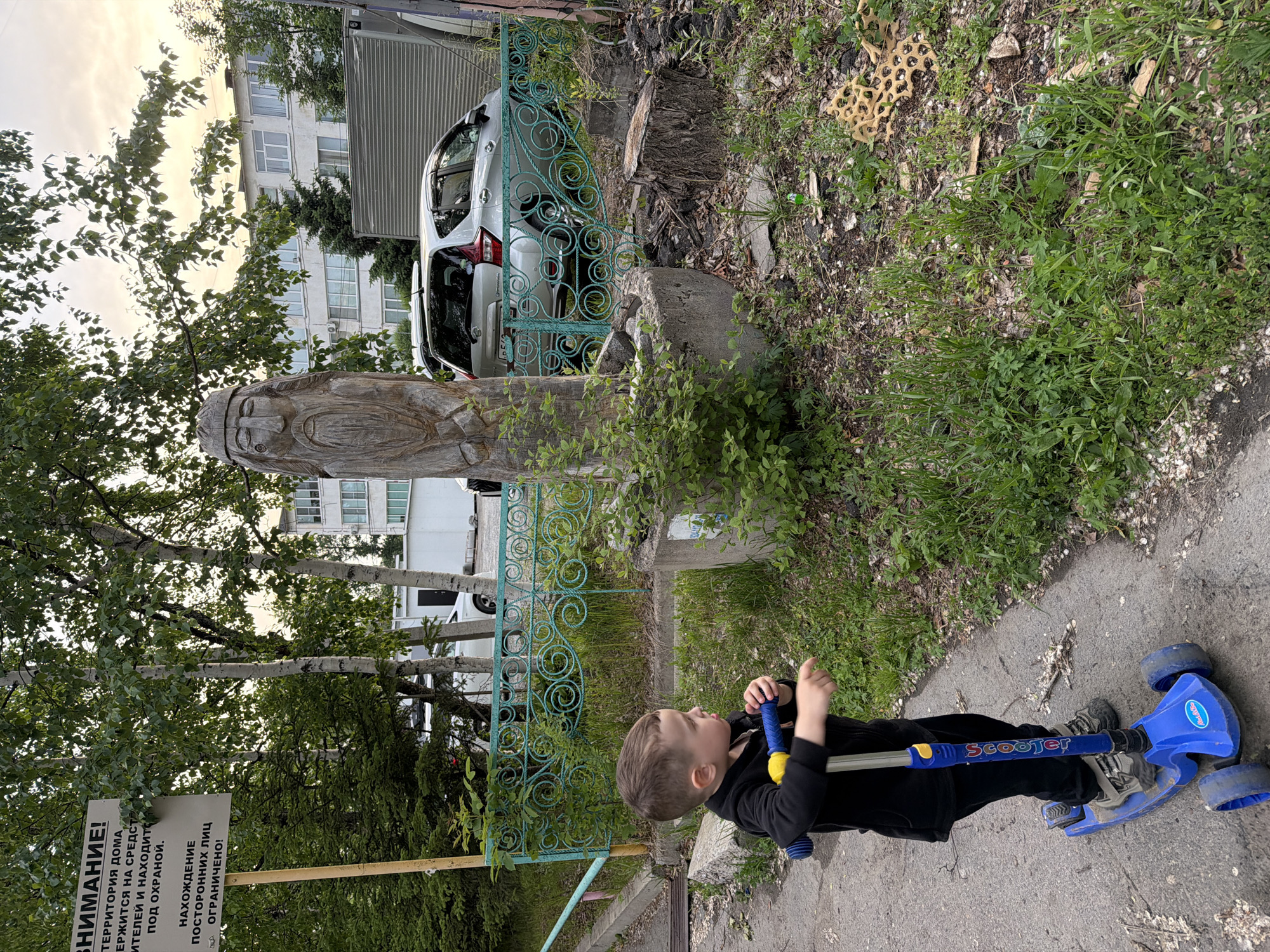A young boy wearing a black zip-up hoodie and black pants is standing on a blue three-wheeled scooter with yellow handlebars. He is looking to his left, with his right hand resting on the scooter's handlebars and his left hand extended as if he is about to push off. The scooter has the word "Scooter" written in blue letters on its stem. He is on an asphalt path that is cracked and has some debris on it. To his right is a grassy area with a weathered wooden sculpture of a face, resembling a folk art piece. Behind the sculpture is a light-colored car parked near a turquoise decorative metal fence. In the background, there are trees, a building with multiple windows, and what appears to be a large gray shipping container. A sign with Russian text is visible on a pole to the left of the image, which reads "ATTENTION! THE TERRITORY OF THE HOUSE IS PROTECTED BY SECURITY SYSTEMS AND IS UNDER PROTECTION. ACCESS FOR UNAUTHORIZED PERSONS IS RESTRICTED!". The scene is outdoors, likely in a residential area of Khabarovsk, Russia, given the language on the sign.