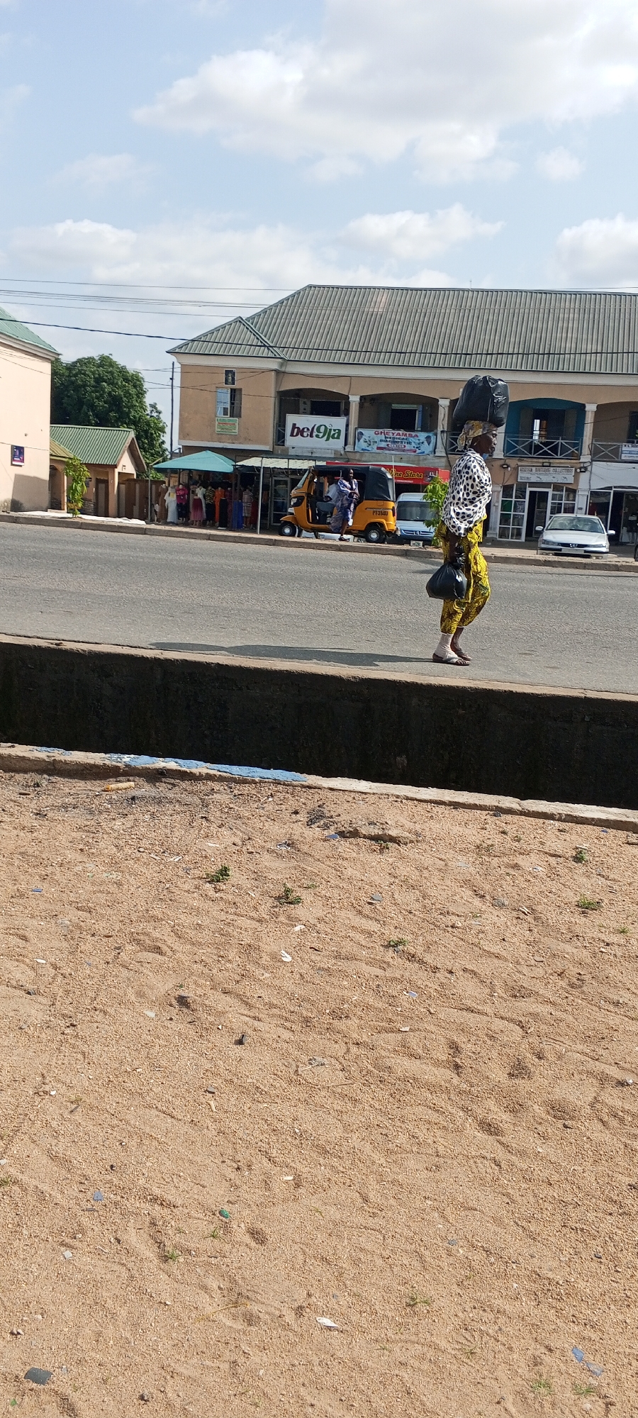 A woman walks on the side of a paved road in Jalingo, Nigeria. She is carrying a large black bag on her head and another black bag in her right hand. She is wearing a patterned white and black blouse and yellow pants with a patterned design. Her feet are bare. In the background, there are buildings, a yellow three-wheeled vehicle, a white van, and a silver car. The sky is partly cloudy. The foreground shows a sandy area with some scattered debris.