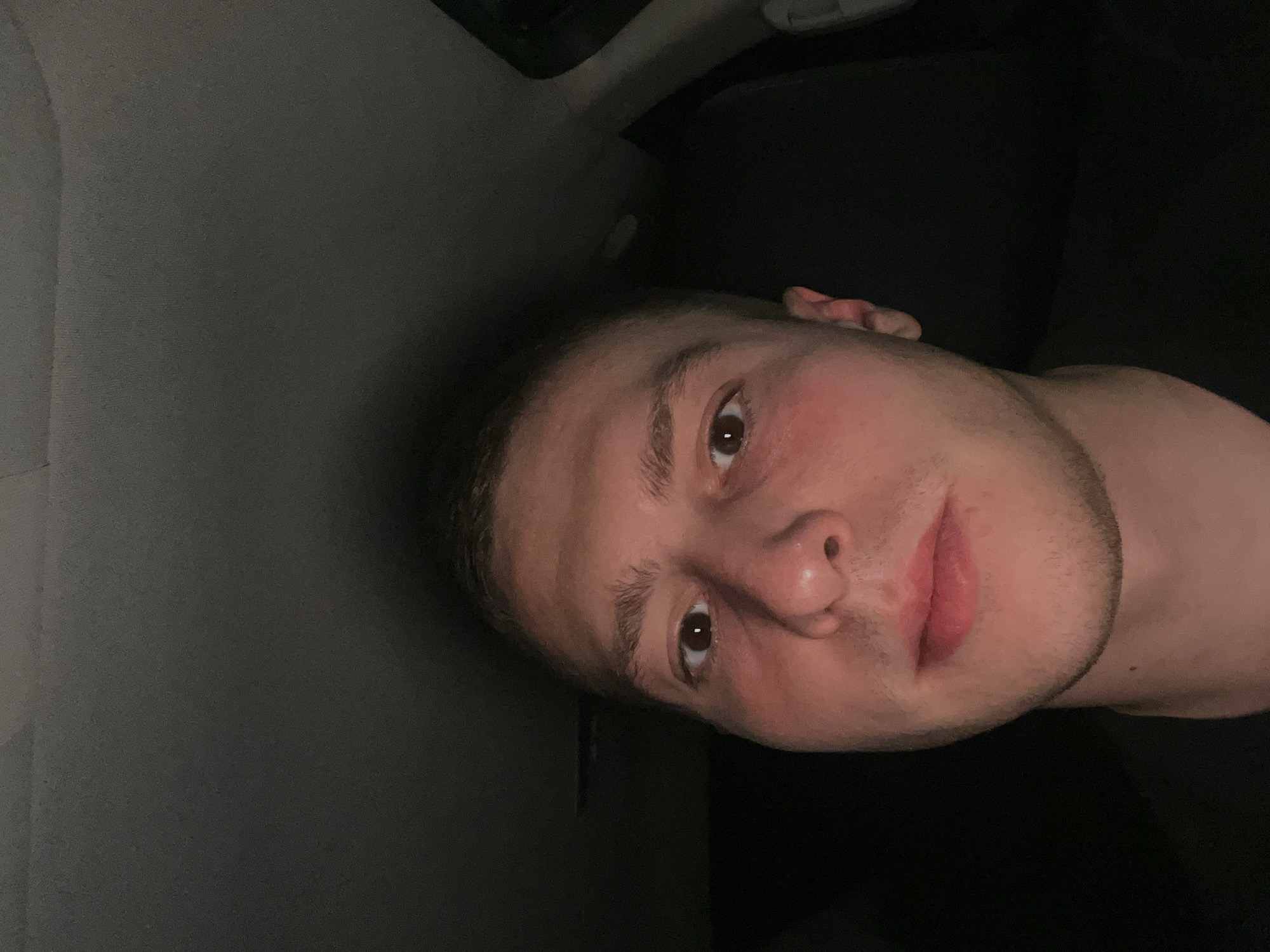 A close-up, slightly rotated image of a light-skinned male is visible, positioned in what appears to be the interior of a vehicle. The subject's face fills the majority of the frame, with details such as brown eyes, short hair, and a slight beard noticeable. The lighting is dim, casting shadows on the face. The background is split between a light grey surface, possibly the car's interior paneling, and a dark area that suggests a seat or the car's ceiling. There's no indication of Burgas, Bulgaria in the image.