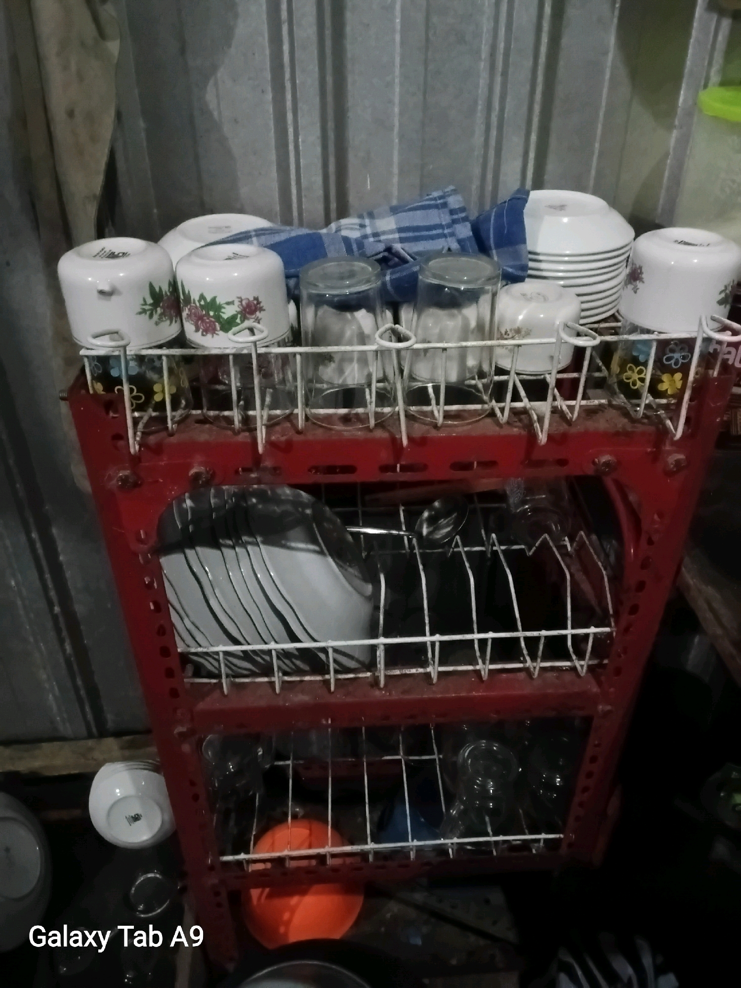 In Bendorejo, Indonesia, a three-tiered red metal dish rack is situated against a corrugated metal wall. The dish rack holds various kitchen items. The top shelf contains white mugs with floral designs, a stack of white plates, and clear glass tumblers. The middle shelf displays a stack of black and white striped plates. The bottom shelf holds several clear glass tumblers and an orange plastic bowl. To the left of the dish rack, a white ceramic bowl sits on the floor, and further left, a portion of a white plate is visible. The lighting is dim, casting shadows. The text "Galaxy Tab A9" is overlaid on the bottom left corner of the image.