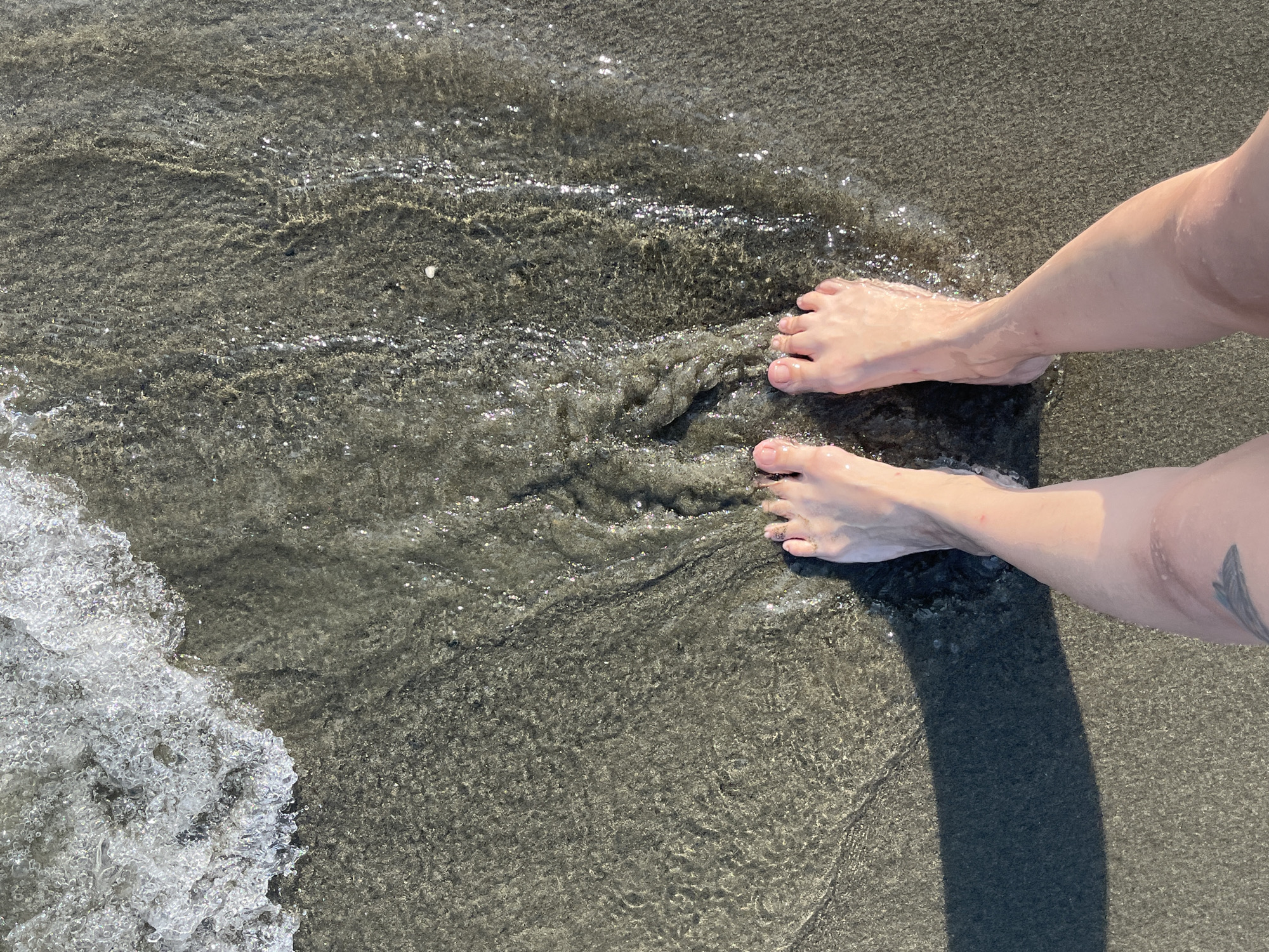 The image shows a person's bare feet standing in shallow water on a sandy beach. The water is clear and gently lapping at the ankles, with a foamy wave visible in the upper left corner. The sand is dark and wet, showing ripples from the water's movement. A shadow of the person's legs is cast on the sand to the left. The scene appears to be in the Agios Tychona Community, Cyprus, a coastal area known for its beaches. The overall impression is one of a peaceful moment at the seaside.