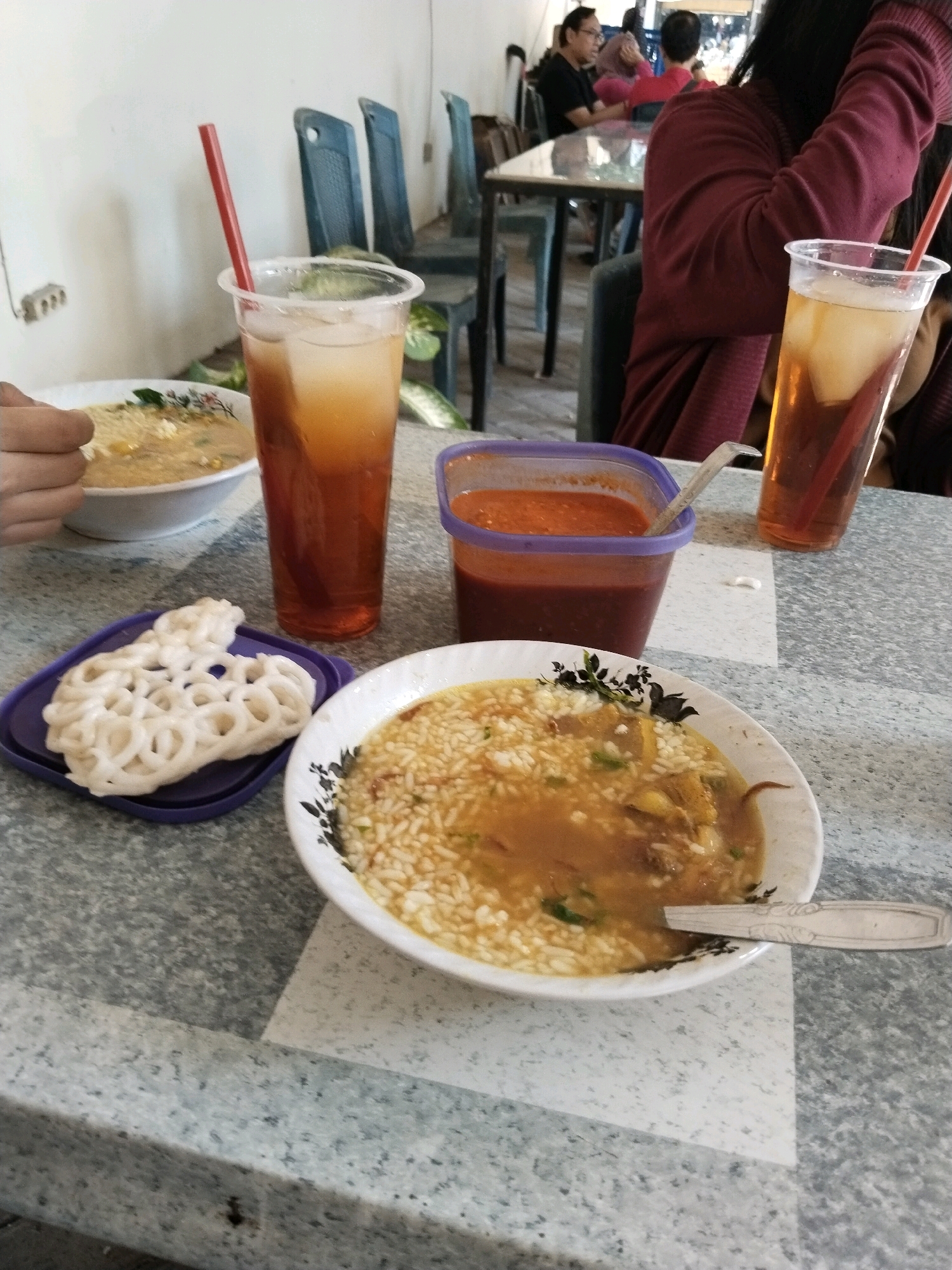 The image depicts a close-up, eye-level view of a table at what appears to be an outdoor food stall or casual eatery, likely located in Surabaya, Indonesia, judging by the context of the food presentation. In the foreground, a white bowl of soup, possibly Soto Ayam, is prominently displayed, with rice, broth, and meat visible. To the left of the soup, a purple coaster holds a white, irregularly shaped kerupuk (cracker). Two tall plastic cups filled with iced tea are positioned behind the soup. One cup, with a red straw, is partially obscured by a hand on the left. The other cup is further to the right. A plastic container of red chili sauce sits between the two cups. In the background, out-of-focus, are several chairs and tables, as well as patrons dining, some of whom are seated and appear to be engaged in conversation. The table surface has a speckled grey and white pattern. The overall impression is of a lively, informal dining scene, characteristic of street food culture in Surabaya.