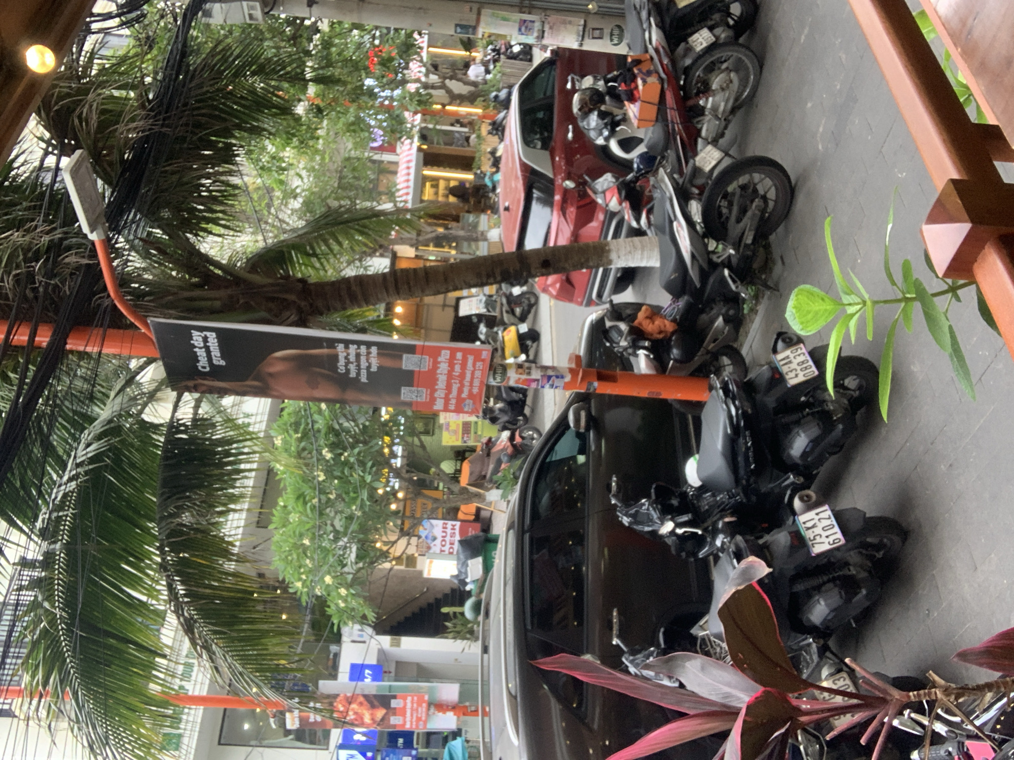 Here's a detailed description of the image:
**Overview:**
The image presents a busy street scene, likely taken in Đà Nẵng, Vietnam. A mix of vehicles, including cars and numerous motorcycles, are parked along the side of the road. Pedestrians can be seen in the background, suggesting a commercial area. The perspective is unusual, as if taken from an elevated vantage point.
**Foreground:**
* Foreground elements include vegetation, possibly potted plants, obscuring the lower right corner.
* Motorcycles are parked densely along the edge of the road. The license plates of some are visible.
**Midground:**
* A red car is parked amidst the motorcycles.
* A black SUV.
* A large banner or sign is prominently displayed, advertising Detroit-Style Pizza. The sign also mentions "Motor City Detroit-Style Pizza," "44 An Thương 3 / 4 pm-1 am," and includes phone numbers. A slogan visible is "Cheat day granted"
* A "TOUR DESK" sign is visible, indicating tourism-related businesses in the area.
**Background:**
* The background depicts a commercial street with various shops and businesses.
* People are walking along the street, indicating a moderately busy environment.
* Trees and other vegetation are visible.
**Environment and Location Clues:**
* The presence of palm trees, typical street signs, and the style of architecture suggest a Southeast Asian location, specifically Đà Nẵng, Vietnam.
**Overall Impression:**
The scene conveys a sense of urban activity in a Southeast Asian city with a mix of local businesses and tourism-related services. The high density of motorcycles parked on the street and the presence of a Detroit-Style pizza restaurant are distinctive features.