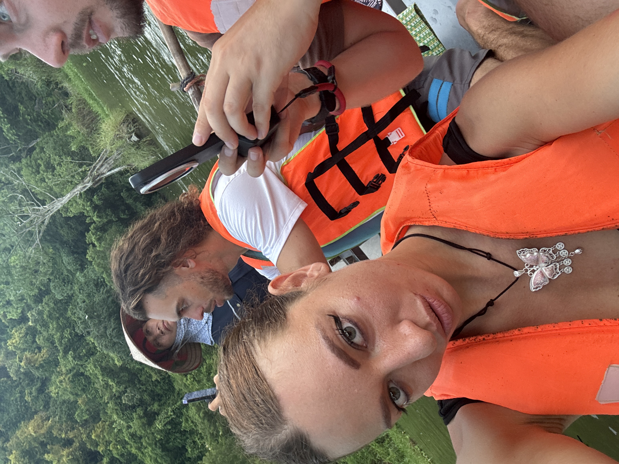 The image captures a group of people on a boat, likely a tourist excursion, in Yên Sơn Ward, Vietnam. In the foreground, a woman is taking a selfie, wearing an orange life jacket and a butterfly pendant. Her hair is pulled back, and she has makeup on, including winged eyeliner. Behind her, a man with long, wavy hair, also wearing an orange life jacket, is holding a smartphone. To the left, a person wearing a conical hat and a light blue headscarf is visible. The background consists of lush green trees and a body of water. The lighting suggests it is daytime.