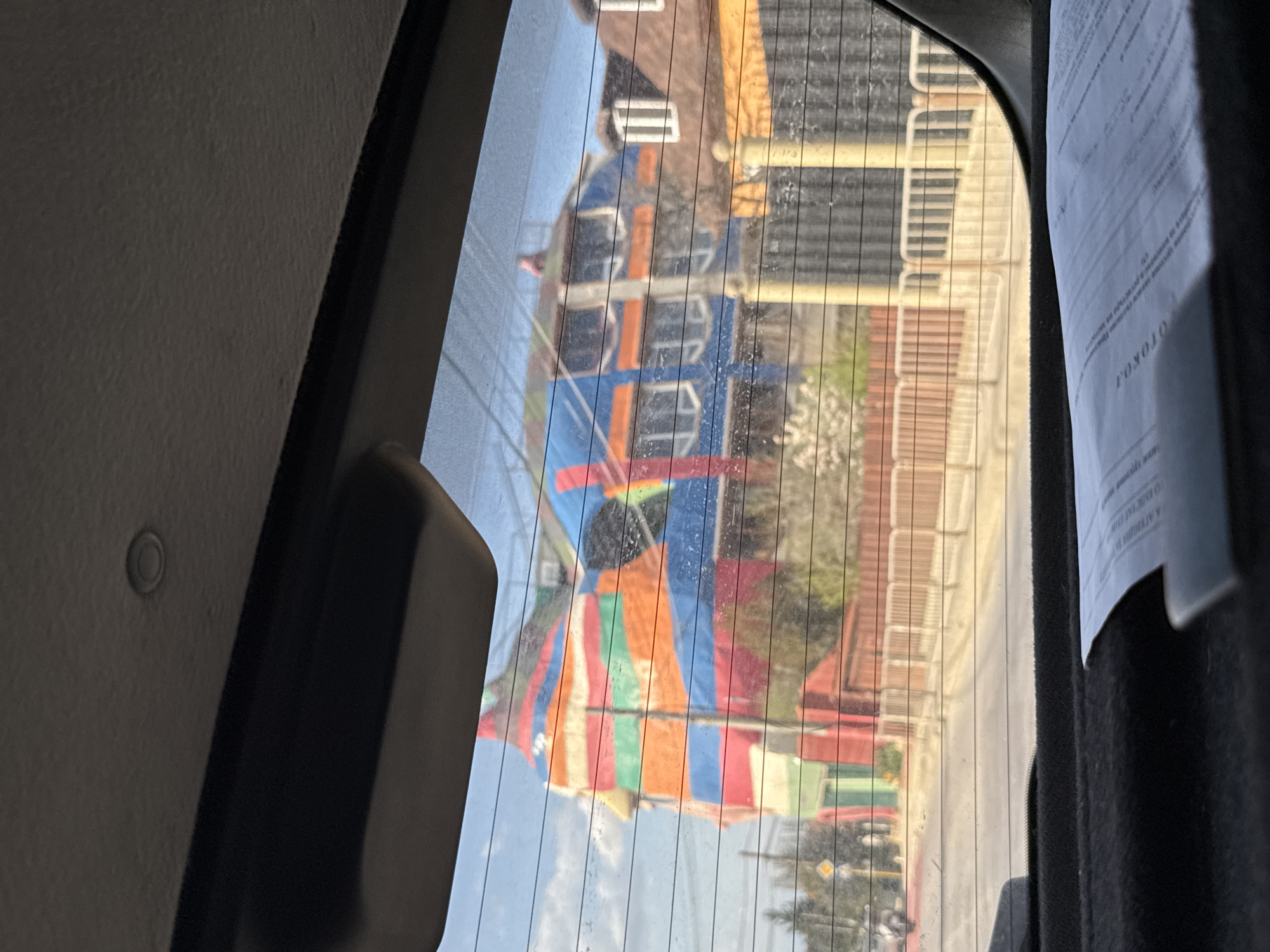 The image presents a view from inside a vehicle, looking out through its rear window. The window features distinct horizontal defroster lines. On the left, parts of the car's interior trim are visible, including a dark panel and a lighter-colored headliner with a small, circular sensor embedded in it. On the right, a white paper document, displaying Cyrillic text, hangs partially obscured; the word "ОТОКО" is clearly visible, reversed.
Outside the window, a bright, sunny daytime scene unfolds. The most prominent feature is a large, colorful structure, resembling an inflatable bounce house or a section of a funfair tent. It is adorned with bold, vertical and horizontal stripes in various hues of blue, orange, red, green, and white. This structure is situated within what appears to be an urban or semi-urban environment.
In the background, several buildings are visible. One building behind the colorful attraction features a brown roof and a lighter facade with multiple windows. Further right, another building displays several levels of white or light-colored balcony railings. Green foliage from trees or bushes is scattered throughout the scene, suggesting natural elements within the built environment. A paved area, possibly a road or pedestrian path, is discernible towards the bottom. The bright illumination indicates clear weather. The Cyrillic text on the document is consistent with the provided location context of Sofia, Bulgaria. No specific activities or individuals are clearly depicted.