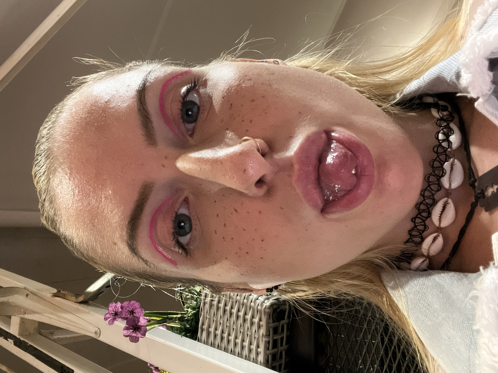 A close-up, low-angle shot depicts a young woman with fair skin, freckles, and blonde hair. She is wearing pink eyeliner and a choker with seashells. Her tongue is sticking out. The background shows a white railing, purple flowers, and a wicker basket. No specific location in Plovdiv, Bulgaria is evident.