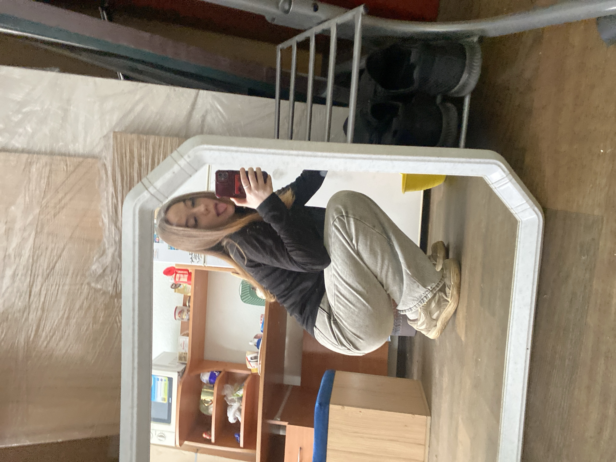 A young woman, approximately 20-30 years old with light brown, shoulder-length hair, is captured in a mirror selfie taken in a room in the city of Chaikovsky, Russia. She is squatting, wearing a black long-sleeved top and light grey jeans, paired with beige sneakers. She holds a dark red smartphone, likely an iPhone, with both hands to take the photograph, her tongue slightly extended.
The background, visible through the mirror's reflection, displays a functional and somewhat cluttered indoor environment. To the left, a light brown wooden desk or shelving unit holds a white microwave oven with an illuminated digital display. Various items are visible on the shelves, including mugs (one gold-colored), bottles, plastic bags, and small containers. Below the desk, a wooden stool with a blue upholstered top is positioned. The floor is light brown, appearing to be wood or laminate.
The wall behind the woman and the desk is covered with large, rectangular items wrapped in clear plastic film, suggesting stored furniture components, insulation, or other packaged goods. To the right of the woman, a silver-colored metal shoe rack is visible, holding at least two pairs of dark-colored athletic shoes on its lower shelf. A bright yellow plastic bucket is on the floor near the shoe rack. More items wrapped in plastic film are visible in the background to the right, along with vertical panels, possibly indicating a storage area or a room in transition.
