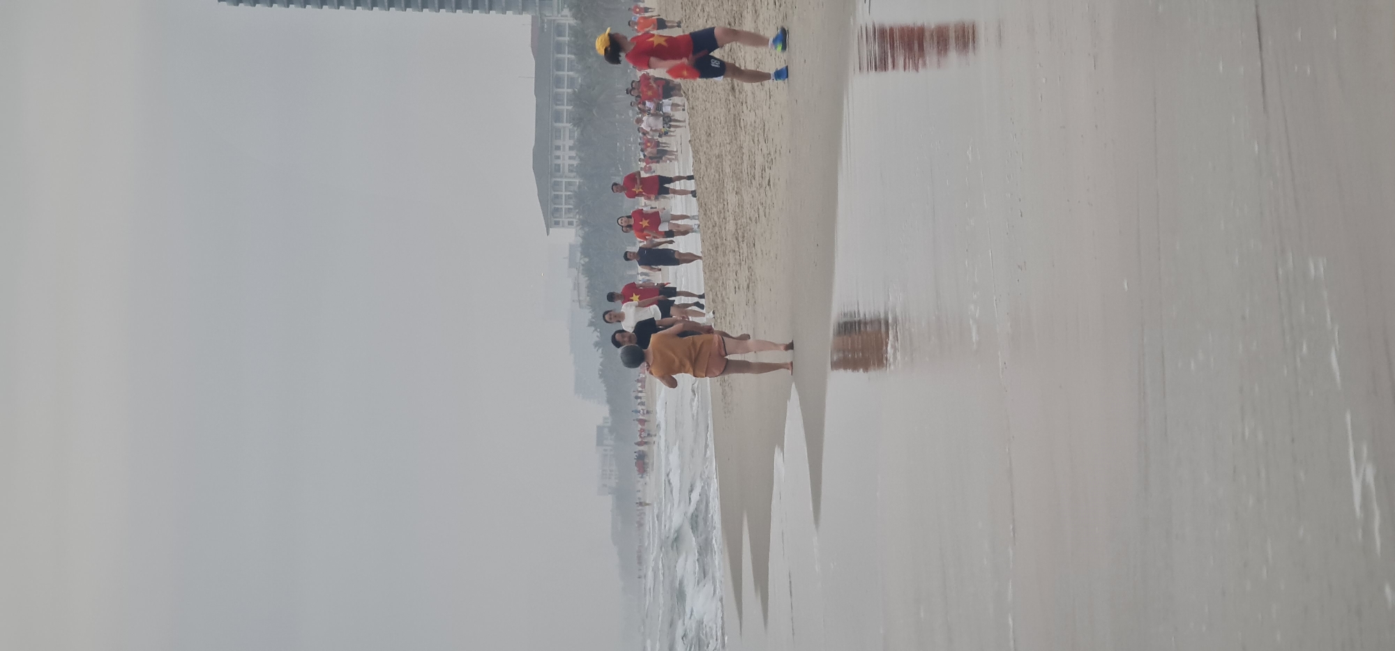 The image depicts a large group of people gathered on a sandy beach under a hazy, overcast sky. Based on the provided context, the location is Da Nang, Vietnam. Many individuals in the group are wearing matching red t-shirts adorned with a prominent yellow five-pointed star, symbolizing the flag of Vietnam, indicating an organized event or group activity.
In the immediate foreground, a person in an orange shirt and dark shorts is seen bending over, possibly stretching or engaging in an exercise. To their right, another individual in a red star-emblazoned shirt, blue shorts with the number "81" visible on them, and a yellow cap is running along the wet sand. The group extends further into the background along the shoreline, with numerous other participants visible.
The beach features wide expanses of wet sand, creating clear reflections of the people and the distant buildings. Gentle waves are breaking on the shore to the left. In the background, a line of multi-story buildings, likely hotels or resorts, can be seen along the coast, somewhat obscured by the atmospheric haze. The scene suggests a daytime event, possibly a morning run or group exercise, against a coastal backdrop.