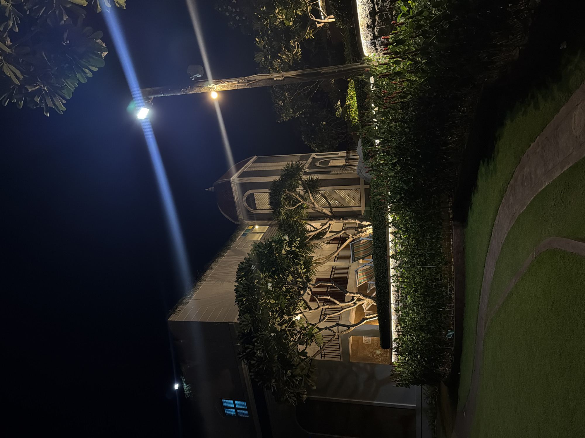 The image depicts a nighttime scene in Bac Nha Trang Ward, Vietnam. A multi-story building is partially visible, illuminated by artificial lighting. The building features a tower-like structure with a red-domed roof. Trees and shrubs are present in the foreground, and a paved pathway winds through a grassy area. A lamppost with two lights casts light onto the scene. The sky is dark.