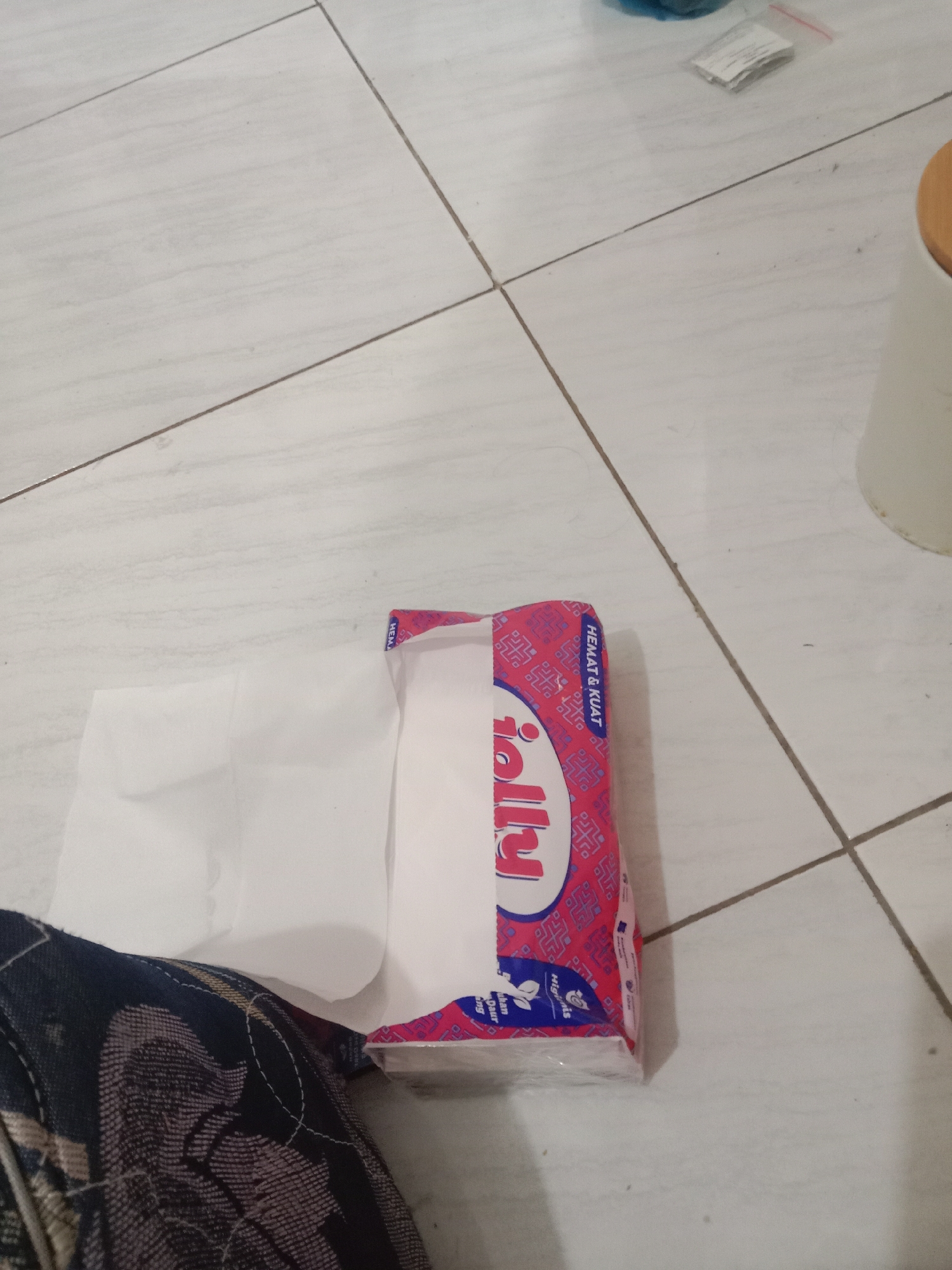The image shows a package of white tissues on a light-colored tiled floor. The packaging is predominantly pink and blue with some white text and graphics. The word "Jolly" is clearly visible in large, white lettering on the front of the package. The tissues themselves are white and appear to be folded. A portion of dark-colored fabric with a patterned design is visible in the lower left corner of the image, partially obscuring the tissues. The tiled floor has thin, dark grout lines. The scene is indoors, and the lighting appears to be artificial, casting a slight shadow from the tissue package. There is no indication of the specific location within Brangsong, Indonesia, beyond the general setting.