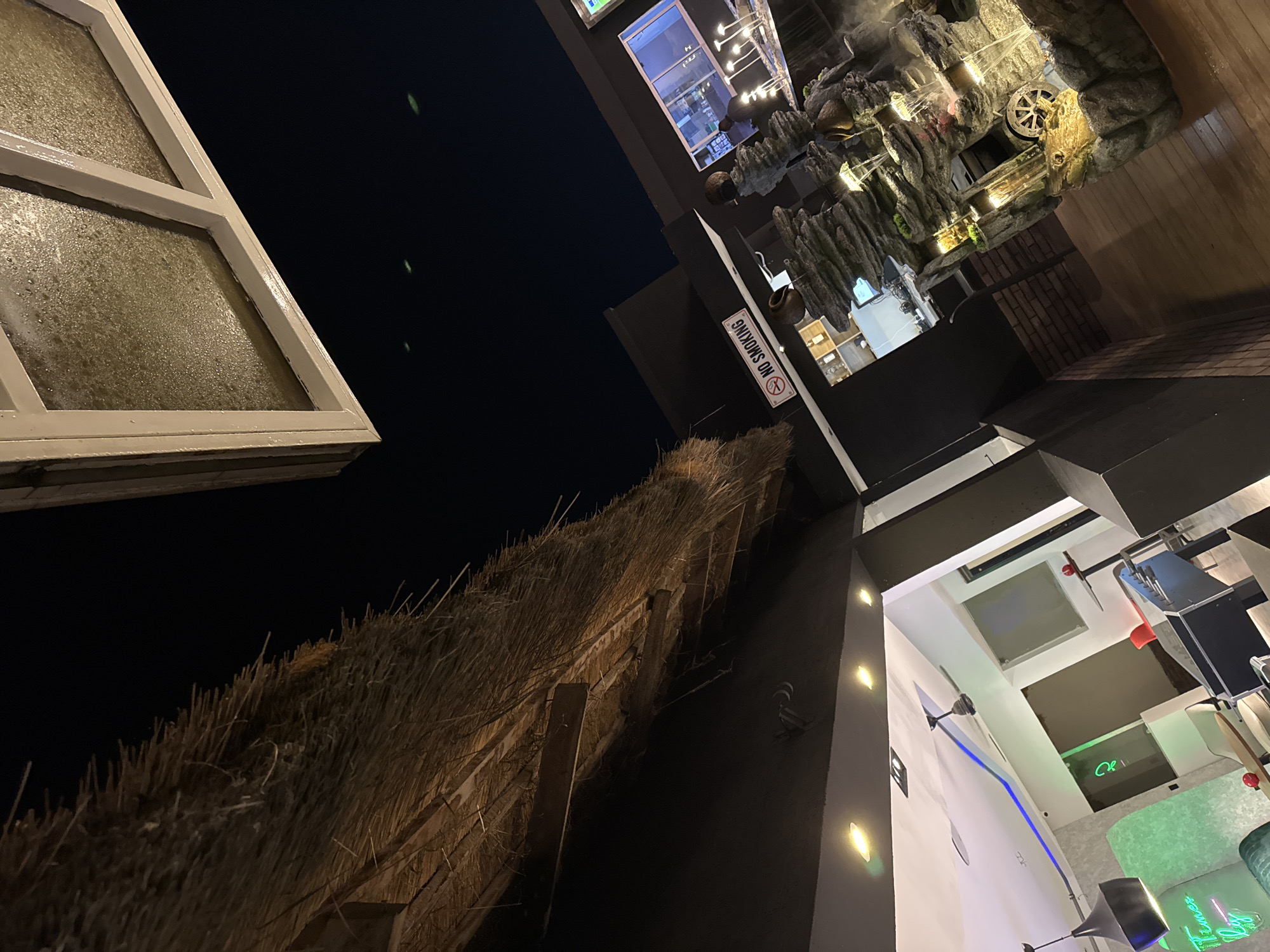 Captured at night in an unknown urban location, the image presents an unconventional angle of various architectural elements. A white-framed window with a textured, possibly water-spotted pane is prominently featured on the left. Adjacent to this is a thatched roof, displaying an aged and organic texture. Signs prohibiting smoking are visible against a dark wall. In the upper right, there's an indoor artificial rock formation with an embedded water wheel. Contemporary interior design elements can be seen in another part of the frame, including ceiling fixtures and lighting installations. The overall scene presents a juxtaposition of traditional and modern designs.