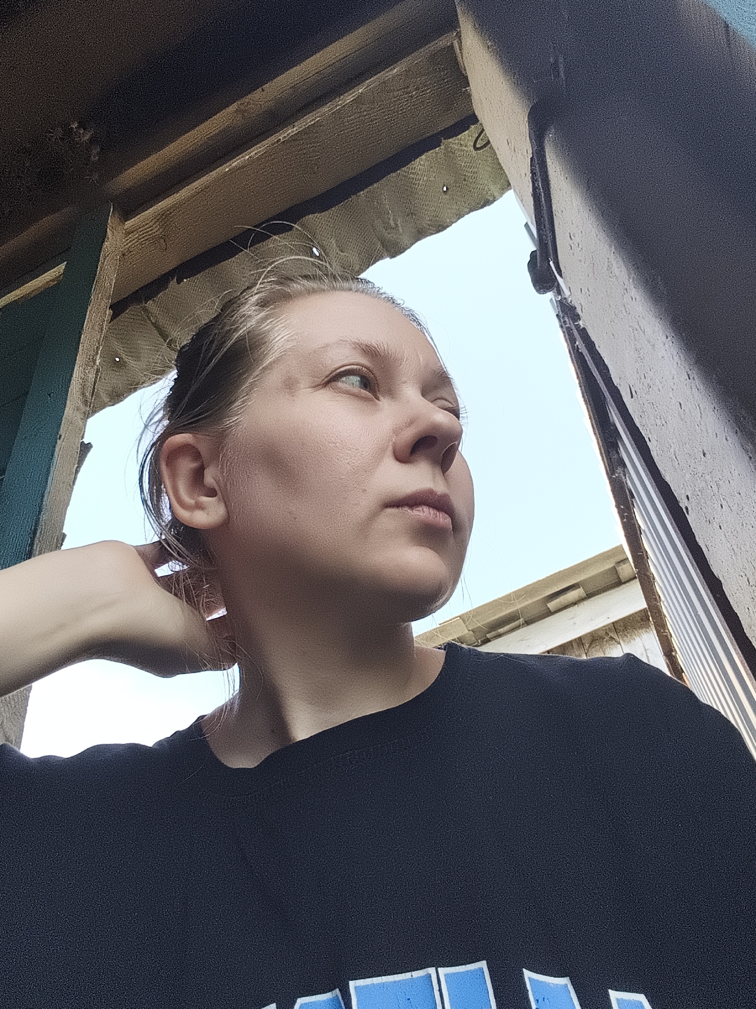 A young woman is captured in a low-angle, close-up shot, looking upwards and to her right. She has light-colored hair pulled back, and her face is illuminated by natural light. Her left hand is raised to her neck, with her fingers touching the back of her head. She is wearing a dark t-shirt. The background consists of a bright blue sky and the weathered wooden structure of what appears to be an outdoor shed or shelter. The structure has visible beams and insulation. The image seems to have been taken outdoors in the village of Ilmino, Russia, given the architectural style and natural light.