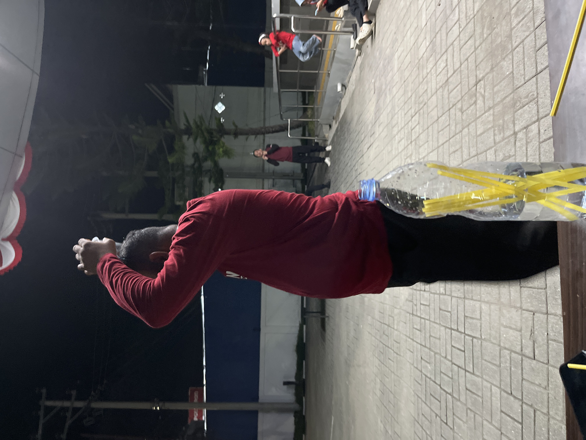 The image depicts an outdoor scene at night, possibly in a public area such as a park or plaza in Kel. Pasangkayu, Indonesia. In the foreground, a person is partially visible from the side, wearing a long-sleeved red shirt and black pants, with their arms raised above their head. To the right of this individual, a large clear plastic bottle filled with water and several yellow straws is prominently placed. The background reveals a paved walkway with a patterned brick design. Several other individuals are visible in the midground and background, including a person in a red shirt and black pants standing further back, and another person in a red top and blue jeans sitting on a railing. The environment includes streetlights, trees, and some structures, suggesting an urban or semi-urban setting at dusk or night.