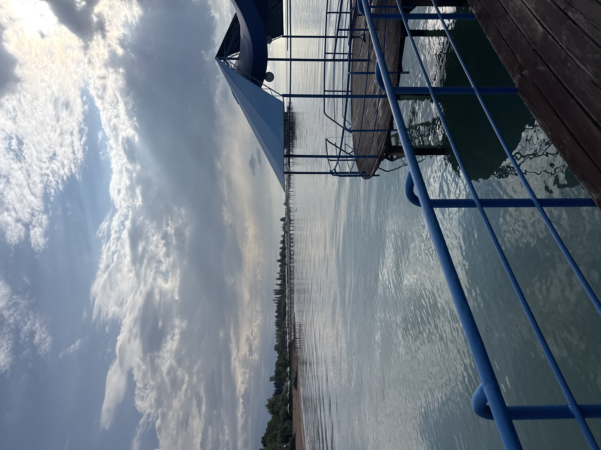 A wide shot captures a serene lake scene in Sary-Oy, Kyrgyzstan, under a partly cloudy sky. In the foreground, a wooden pier with blue railings extends into the calm, reflective water. A covered structure with a blue canopy sits on the pier. The middle ground shows the expanse of the lake, with a distant shoreline lined with trees and buildings. The background features a hazy mountain range beneath a dynamic sky. The overall impression is one of peaceful, natural beauty.