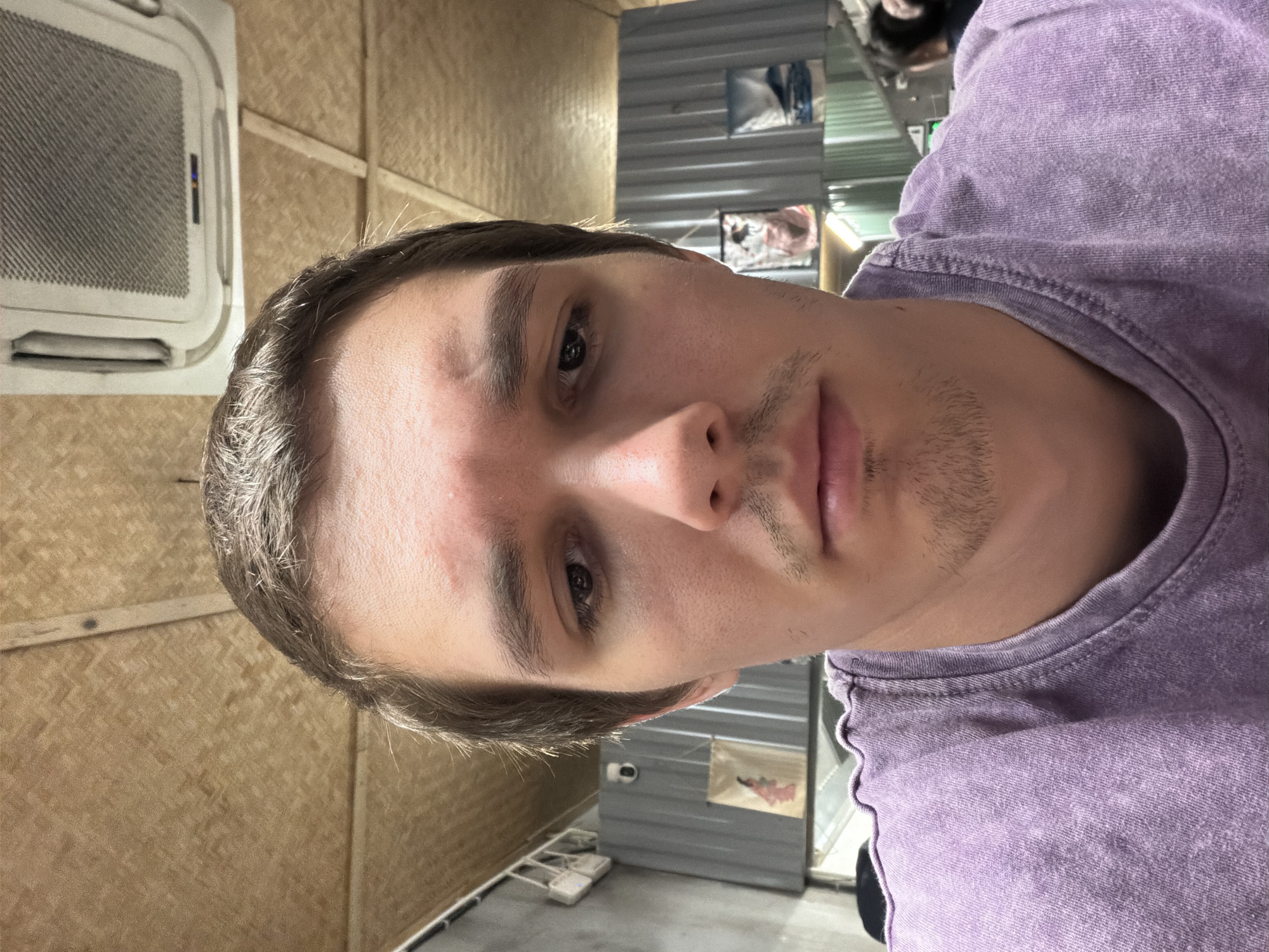 A vertical, eye-level close-up image captures a young man, likely in his late teens or early twenties, looking directly at the camera with a neutral expression. He has short brown hair, a light mustache, and a faint goatee. He is wearing a faded, textured, light purple t-shirt.
The background is divided into two distinct parts. To the left, behind the man's head and left shoulder, is a wall covered in a light brown, woven material, possibly bamboo. A white, rectangular, ceiling-mounted air conditioning unit with a gridded front and a small blue illuminated display panel is visible on this wall.
To the right, behind the man's right shoulder and torso, is a corrugated metal wall or partition. This surface has several reflective areas, showing blurry forms that appear to be other mirrors or indistinct objects. A small security camera is mounted on this corrugated wall, and below it, a small framed picture or notice is attached. The floor in this area is dark, with some small white electrical items or devices visible.
The overall lighting is artificial and somewhat dim, suggesting an indoor setting. No specific time of day or weather can be discerned. There is no visible text within the image. The mix of materials—woven wall, corrugated metal, and a modern AC unit—suggests an informal or utilitarian indoor space, consistent with a location such as Đà Nẵng, Vietnam.