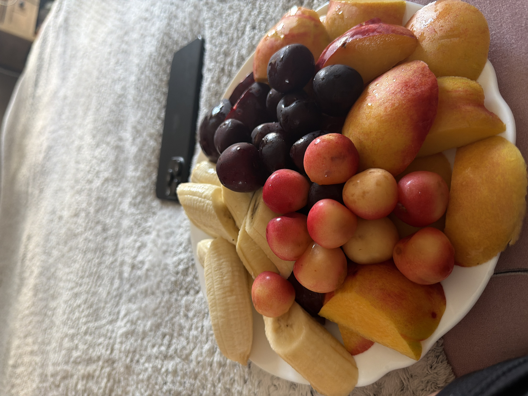 A white plate filled with sliced bananas, peaches, and dark cherries is situated on a textured, light-colored surface, possibly a bedspread. A dark-colored smartphone is partially visible in the background, resting on the same surface. The scene appears to be indoors, likely in a domestic setting. There are no people or animals visible in the image. The lighting suggests it might be daytime, with soft light illuminating the fruit. The overall impression is one of a casually arranged snack or meal.