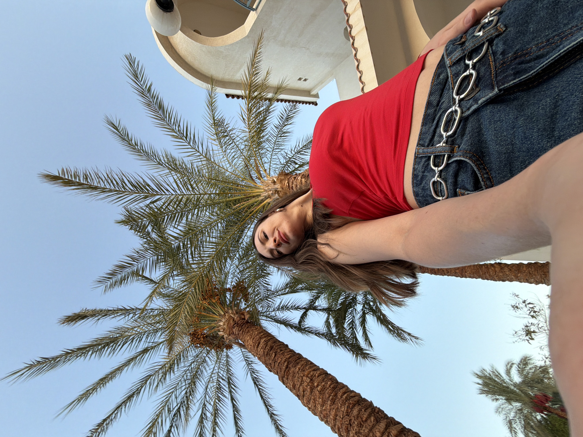 A low-angle, outdoor shot captures a woman posing beneath palm trees in Sharm El Sheikh, Egypt. The woman, with long brown hair, wears a red crop top and denim jeans adorned with a silver chain belt. She looks upwards towards the camera, with a slight smile. Behind her, the rough bark of a large palm tree is visible, its fronds reaching towards a clear blue sky. To the right, a section of a light-colored building with a rounded archway and an outdoor light fixture is present. The scene is bathed in natural daylight, suggesting it is daytime.