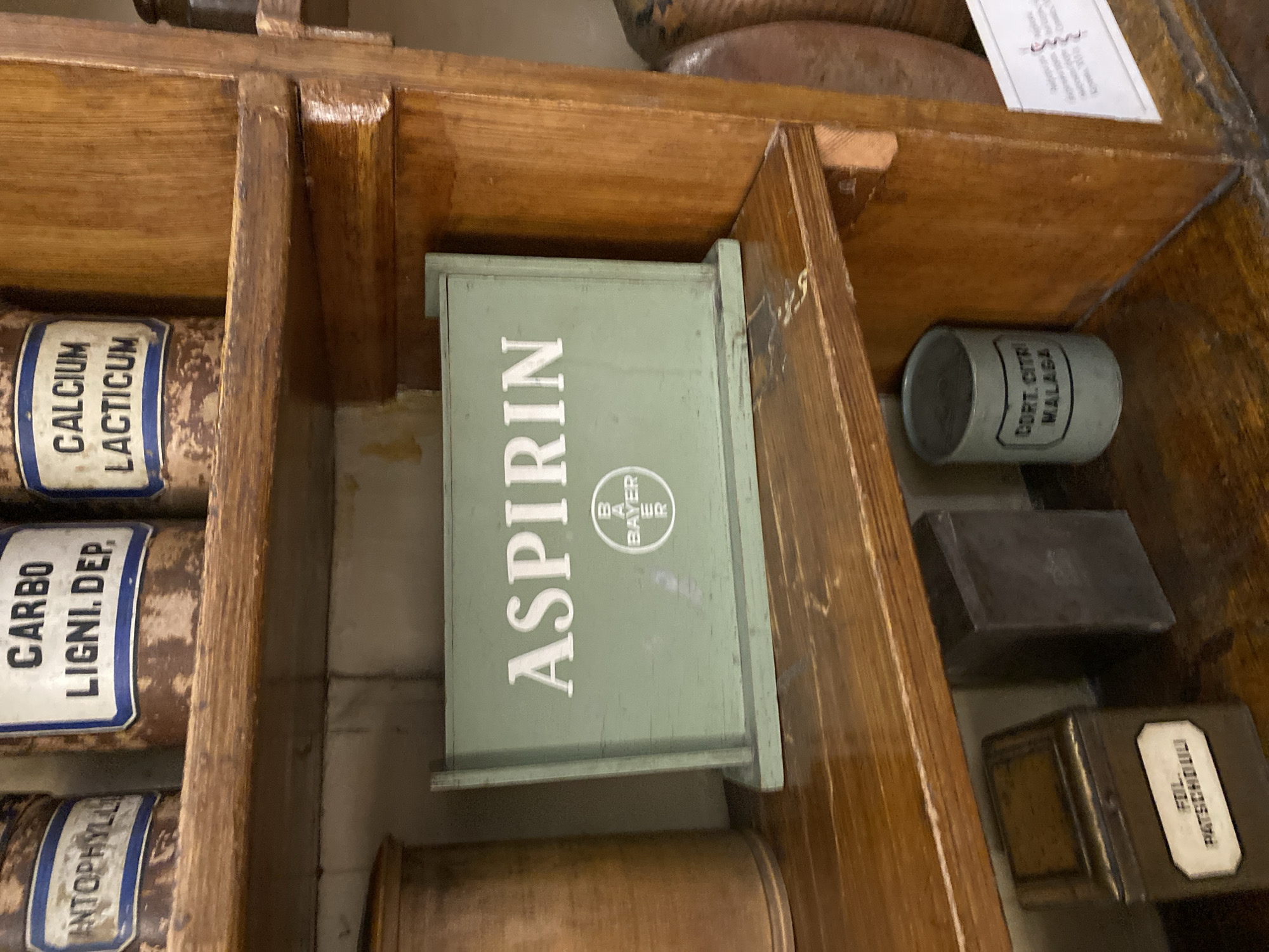 The image shows a close-up view of shelves in an old apothecary or pharmacy. The shelves are made of wood and appear to be aged. On the top shelf, several old metal canisters with labels are visible. One canister has a label that reads "CARBO LIGNI. DEP." and another "CALCIUM LACTICUM". To the left of these, another label is partially visible, reading "ANTOPHYLLI".
On the shelf below, a prominent light green wooden box is centered, displaying the word "ASPIRIN" in white capital letters. Below "ASPIRIN", the Bayer cross logo is also visible. To the left of the Aspirin box, part of a large wooden canister is visible.
On the shelf below that, a small, rectangular metal tin with a label reading "FIL. PATSCHDULI" is visible on the left. Next to it is a dark, rectangular metal container. On the right side of this shelf, a light blue cylindrical container is visible, with a label reading "CORT. CITRI MALAGA". Behind this, a section of a large, brown, textured object (possibly another canister or vessel) is visible.
A white paper with text and a coiled illustration is affixed to the wooden vertical support on the far right of the image. The overall setting suggests a historical display of medicinal items, likely within a museum or preserved apothecary. The specific location in Lviv, Ukraine, is not discernible from the image itself.