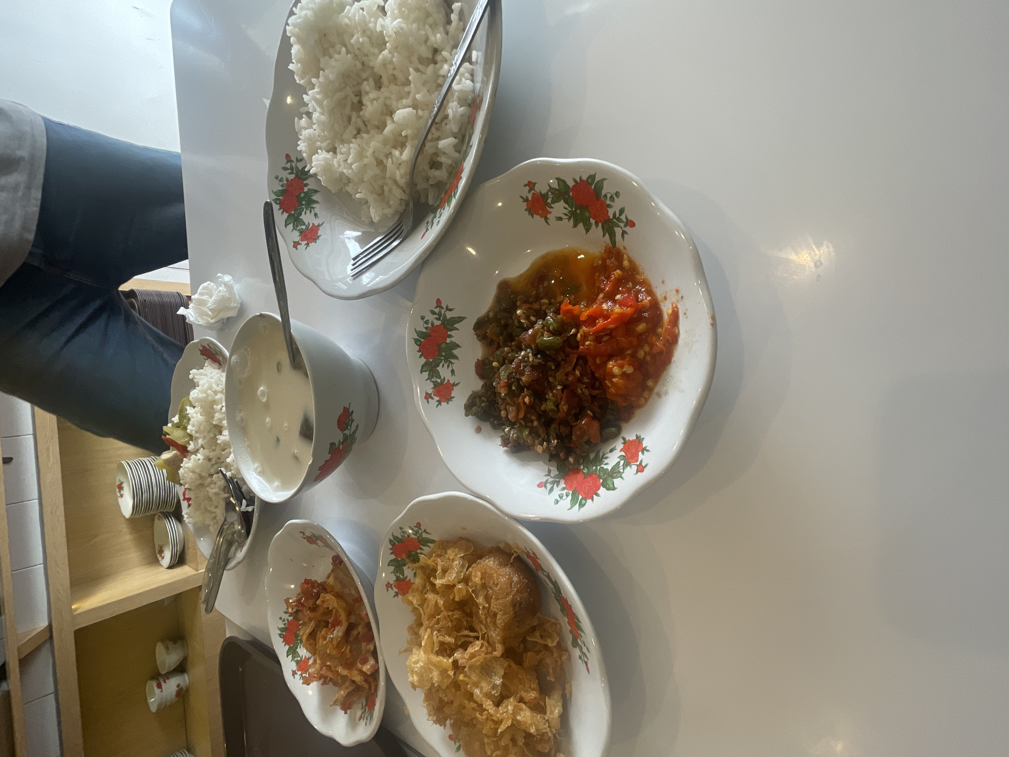 A table setting for one, likely in a casual eatery in Medan, Indonesia, features several small plates of food and a bowl of soup or sauce. The table is white and reflects light. In the foreground, a white, shallow bowl with a floral pattern holds a portion of red chili paste with visible chopped chili peppers and some oil. To the left, another similar bowl contains a fried dish with a crispy, golden-brown batter, possibly a type of fried chicken or seafood. Further left, a smaller oval dish holds what appears to be dried or crispy fried food items. Behind these, a larger white plate holds a generous serving of white rice, with a fork resting on it. A white bowl with a red and green floral pattern contains a pale liquid, possibly soup or a sauce. In the background, a stack of small white plates is visible, along with some decorative cups. A person's legs, clad in blue jeans, are visible in the upper right portion of the image, suggesting they are seated at the table. The overall impression is of a simple meal served in a clean, well-lit environment.