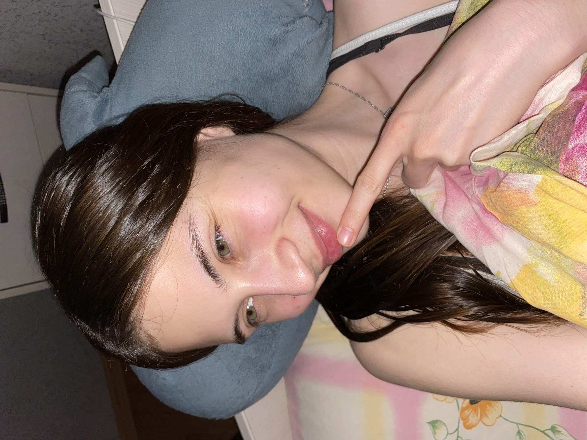 A young woman lies on a bed with a floral patterned blanket. Her head rests on a blue stuffed animal. Her hair is dark brown and shiny. She is touching her lip with her right index finger. A silver chain necklace is visible. No information suggests this image was taken in Votkinsk, Russia.