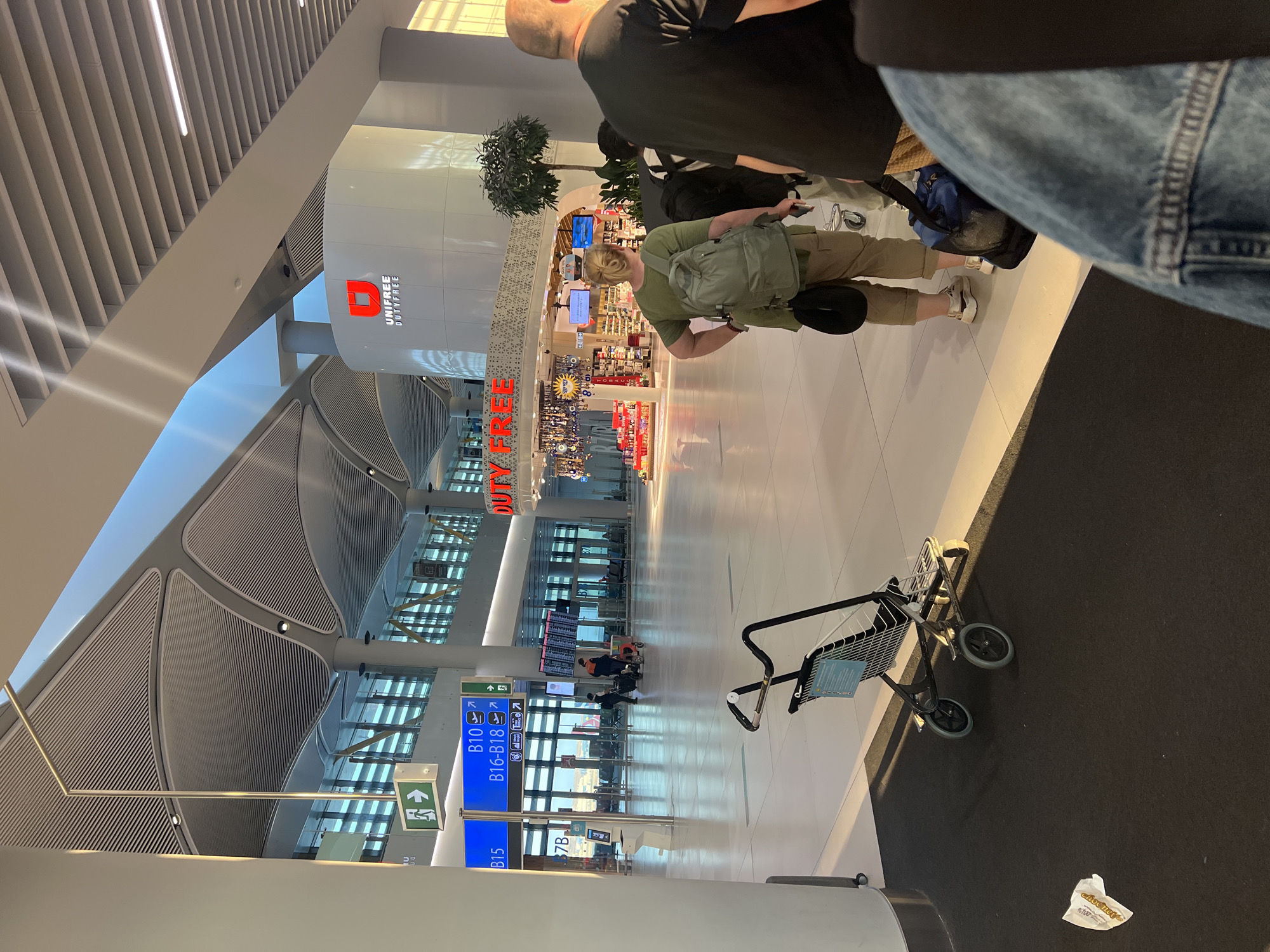 The image depicts the interior of a modern airport terminal, likely Istanbul Airport in Arnavutköy, Turkey, judging by the "Unifree Duty Free" signage. The spacious hall features a high, arched ceiling with integrated lighting and ventilation. Natural light streams in from large windows visible in the background.
In the foreground, a person wearing a green t-shirt and khaki pants, carrying a backpack, is standing with their back to the camera. To their right, another person is partially visible, also facing away. In the middle ground, a luggage trolley is positioned on a darker carpeted area. Further back, an "Unifree Duty Free" store is visible with various products on display. Multiple individuals, some with luggage, are scattered throughout the terminal, walking or standing. Directional signs, including an exit sign and signs indicating gate numbers (B10, B15, B16-B18), are mounted on pillars. The floor is a light-colored, polished surface.