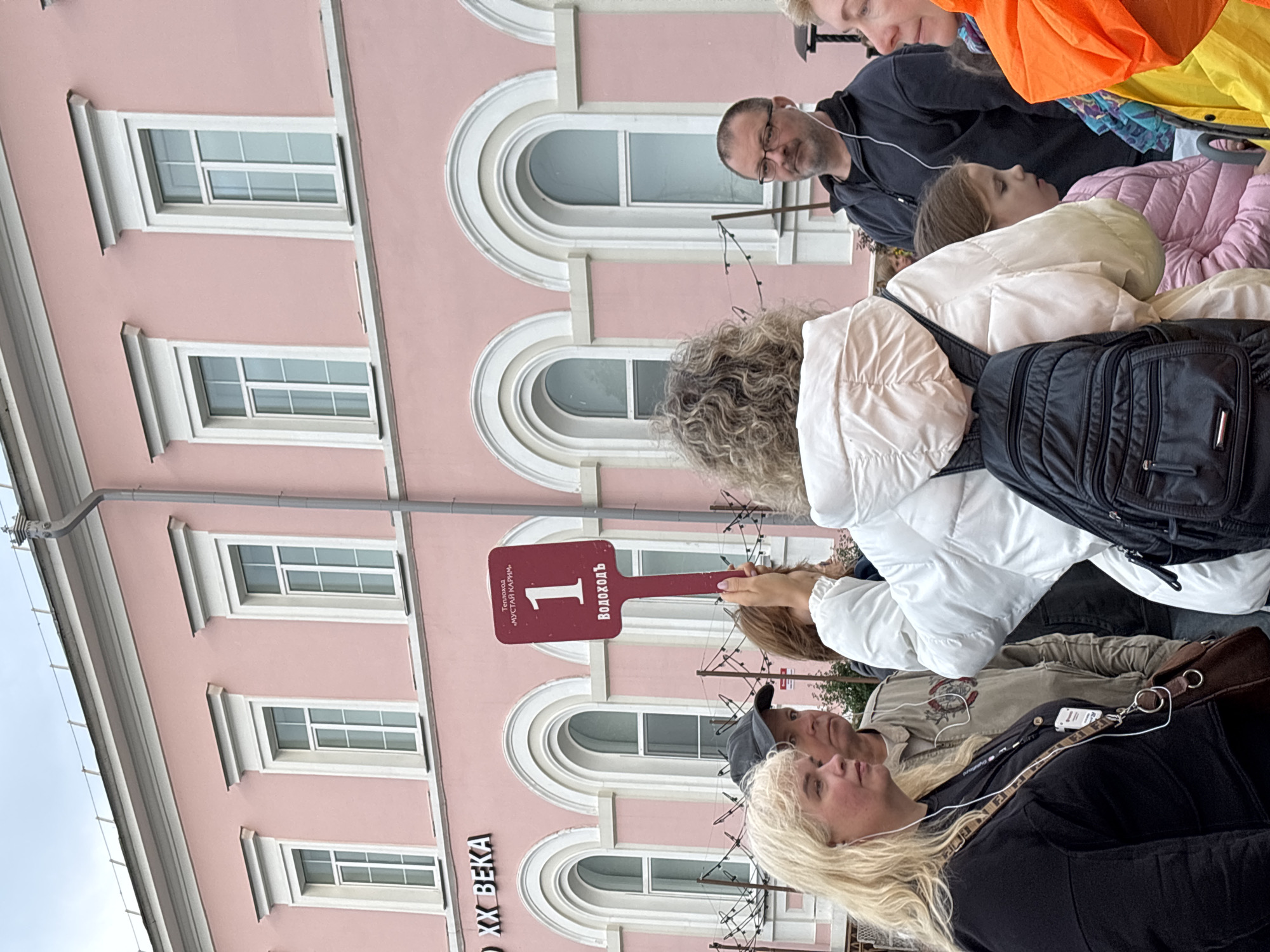 In front of a pink building, several people are gathered outdoors. A woman in the foreground is wearing a black top and has blonde hair. She is looking to her left. To her right, a man with a grey cap is looking forward. Behind them, a woman in a white puffer jacket is holding up a red sign with white text that reads "1" in large font, and "Теплоход «МУСТАЙ КАРИМ»" and "Водоходъ" in smaller font. To the right of the woman holding the sign, a man with glasses and earbuds is looking forward. Further to the right, a young girl in a pink jacket is looking down. The building behind them has arched windows and decorative trim. The ground is paved. The text on the building in the upper left corner reads "XX ВЕКА". This scene is located in Nizhny Novgorod, Russia.