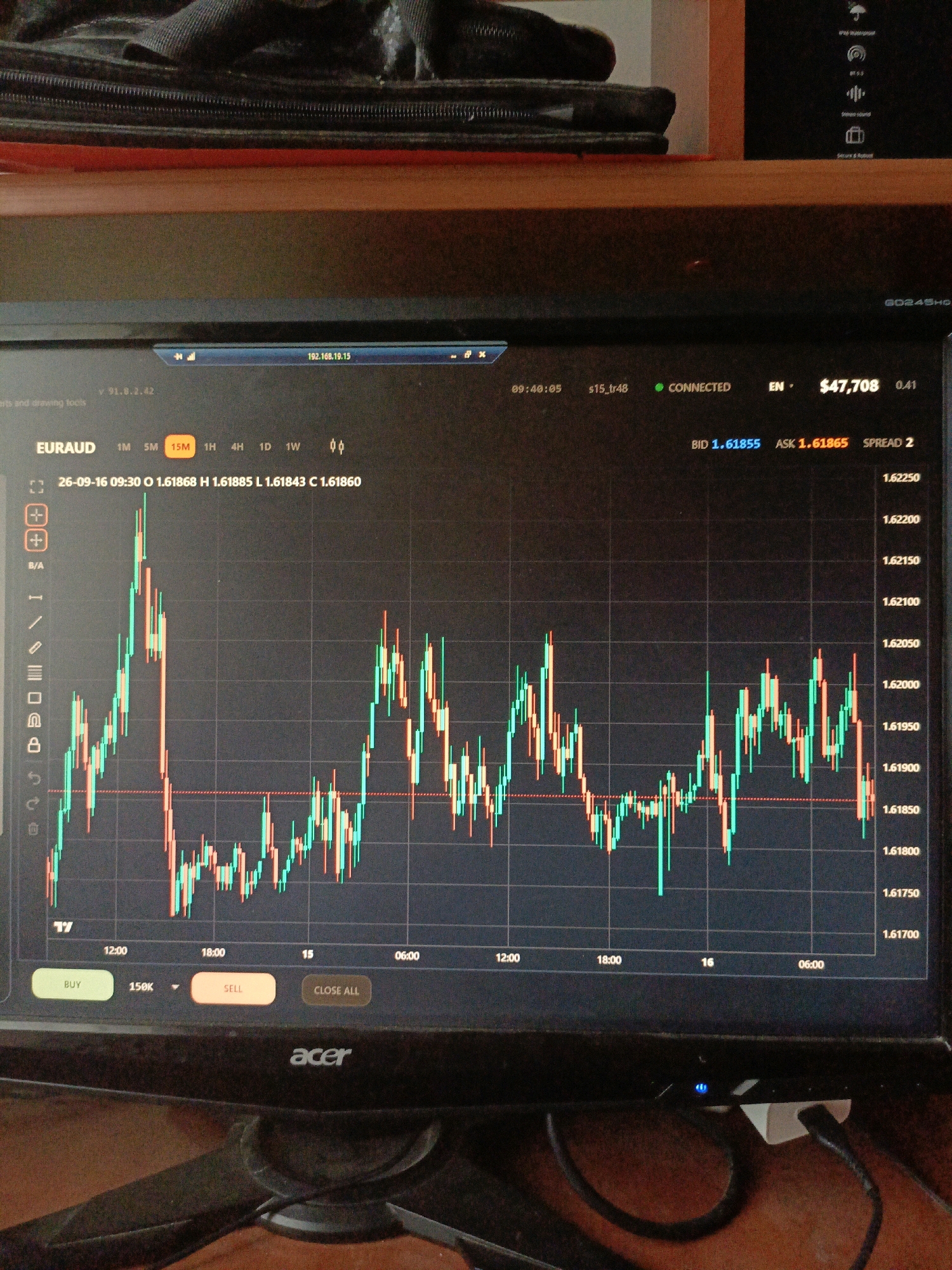 The image shows a computer monitor displaying a financial trading chart. The chart shows the EUR/AUD currency pair with candlestick patterns representing price movements over time. The time frame selected is 15 minutes. The monitor is placed on a wooden surface, and the background shows a dark fabric object. The screen displays various trading information, including account balance, bid and ask prices, and order buttons. The Acer logo is visible on the bottom bezel of the monitor. The scene is indoors and appears to be a home or office setting. There are no people visible in the image. The location of Ухманы, Russia is not discernible from the image.