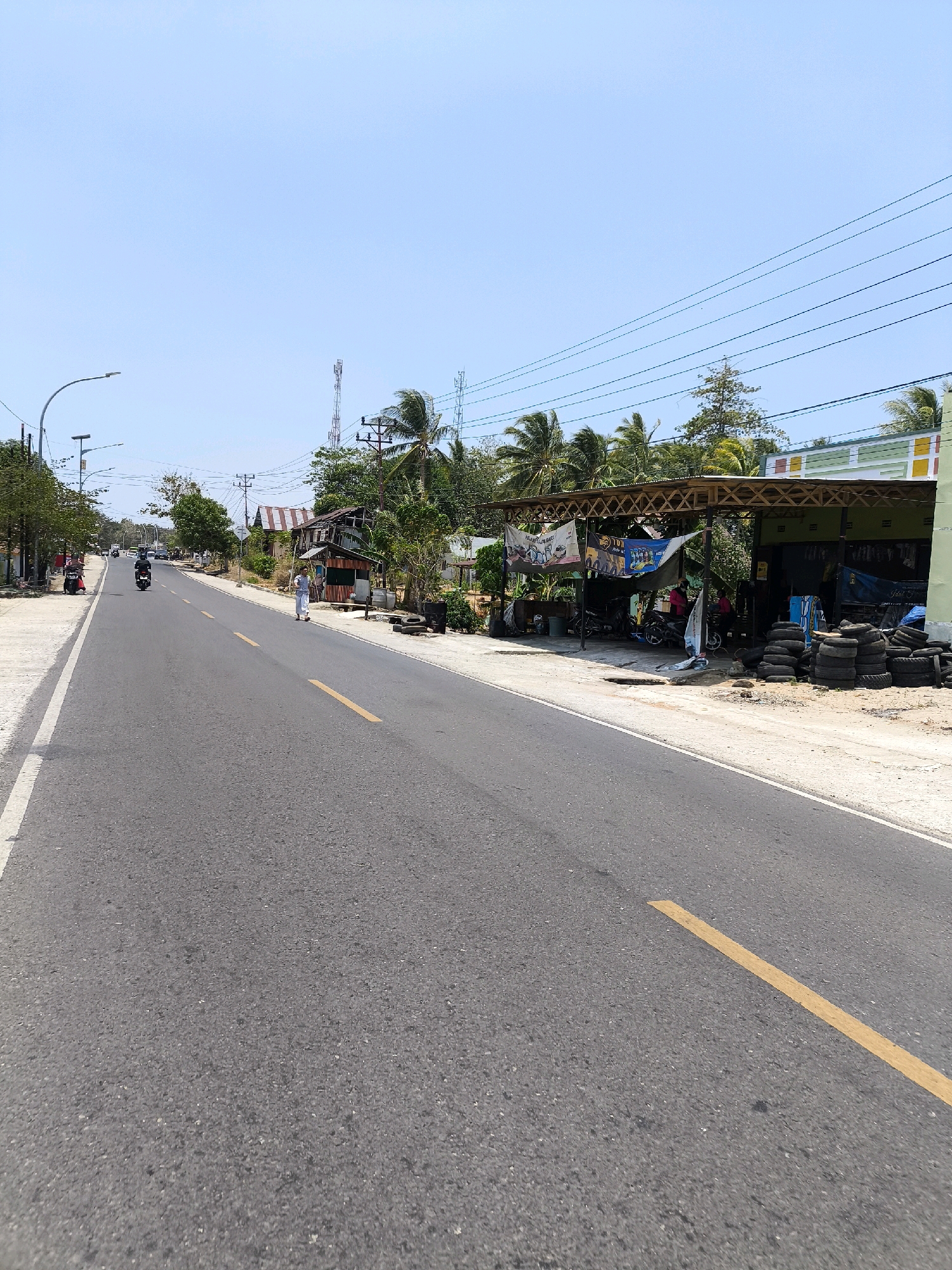 This image captures a street scene in Kwandang, Indonesia, on a clear, sunny day. A paved road runs through the center of the frame, marked with a yellow line down the middle and a white line along the left edge. On the right side of the road, a small establishment with a corrugated metal roof and open sides displays stacked tires. Behind it, buildings with various colored facades and awnings are visible. Lush green palm trees and other foliage frame the scene, interspersed with utility poles and power lines that stretch across the bright blue sky. Several people are present: two individuals are riding motorcycles on the road, and one person is walking on the sidewalk. In the background, more buildings and trees line the street, and a tall telecommunications tower is visible against the horizon. The overall atmosphere suggests a warm, tropical environment with a modest commercial area situated along a main thoroughfare.