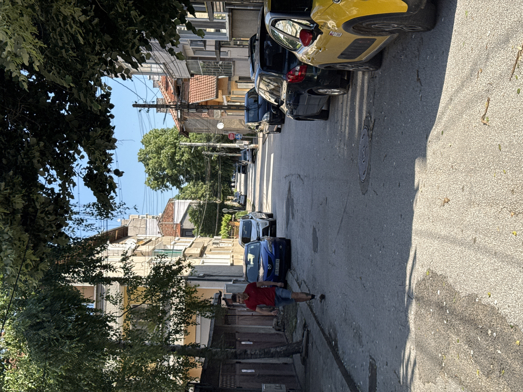 A street scene in Lovech, Bulgaria, is captured in this medium, slightly rotated, outdoor shot. The street stretches into the distance, lined with parked cars on the right and buildings interspersed with trees on the left. A man in a red t-shirt and blue shorts is walking on the left side of the street. Vehicles of various colors (yellow, black, blue) are parked along the right side of the road. Buildings of different heights and architectural styles are visible on either side of the street. Overhead wires crisscross above the street. The sky is visible in the distance.