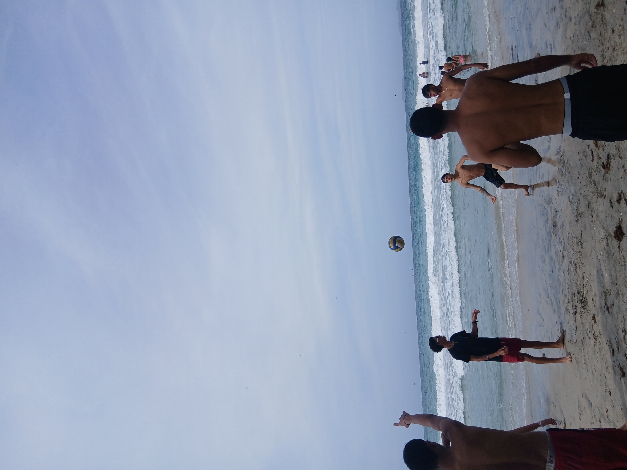 The image depicts a scene at Playa Parguito, Venezuela, where a group of people are playing volleyball on the beach. The players are primarily young men, some shirtless and others wearing t-shirts and shorts. A volleyball is visible mid-air. The beach stretches along the coast with waves breaking along the shore. The sky is light blue, indicating a clear day.