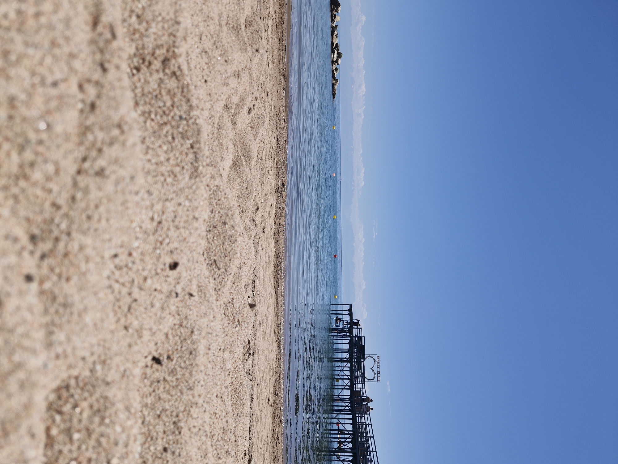 This is a low-angle, ground-level photograph of a beach on a clear, sunny day, looking out towards a body of water and a distant mountain range. In the foreground, the sand is sharply focused, showing individual grains and texture. Beyond the sand is a calm body of water, appearing to be a lake, with a gentle ripple on its surface. Scattered yellow and orange buoys are visible in the water.
To the right of the frame, a wooden pier extends out over the water. Several people are visible on the pier, some standing and others walking. At the end of the pier, a decorative element with the words "FAMILY PLACE" and a heart-shaped frame is mounted. A few individuals are on the pier, and some appear to be interacting or observing the water.
In the distance, a line of rocky formations is present on the left side of the water. Further back, snow-capped mountains are visible on the horizon, under a vast, cloudless blue sky. The scene suggests a recreational area, likely a beach resort.
The location is identified as Bulan-Segettu, Kyrgyzstan, a place known for its beaches along Issyk-Kul Lake, which is consistent with the features depicted in the photograph.