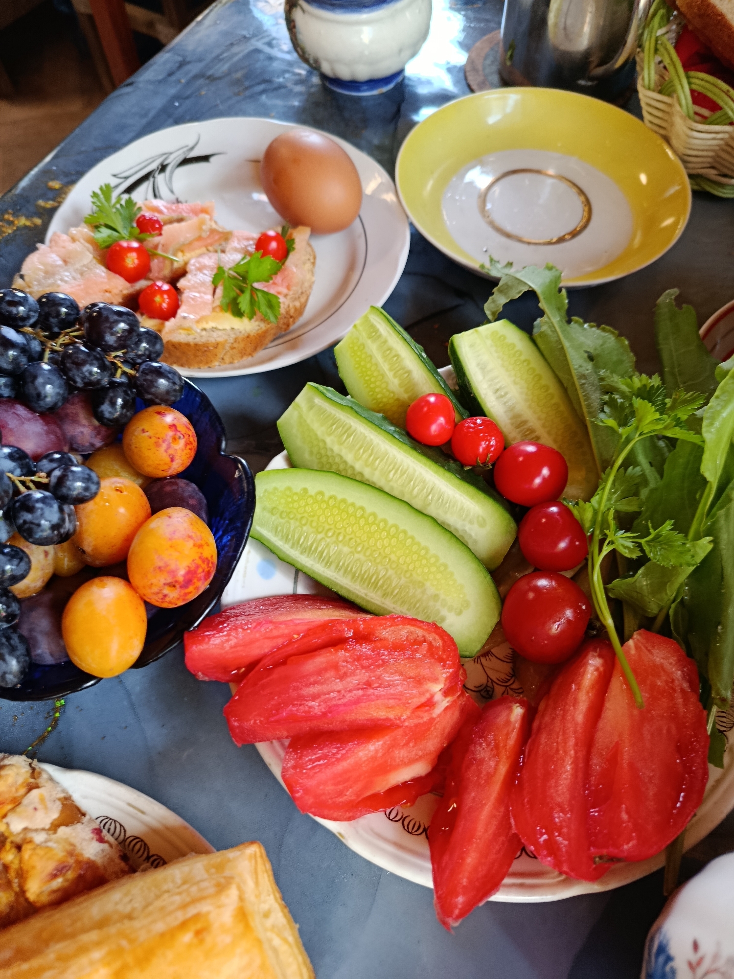 A table is set with a breakfast or light meal, featuring several dishes. In the foreground, a plate holds sliced tomatoes and greens. To the left, a blue bowl contains a mix of dark grapes and small orange plums. Behind the fruit, a white plate displays a hard-boiled egg and open-faced sandwiches topped with slices of smoked salmon, cherry tomatoes, and parsley. Another white plate holds more cut tomatoes and greens. A yellow and white bowl sits further back. The scene appears to be indoors, with a dark tabletop. The image context suggests this meal is taking place in a home in Verea, Russia.