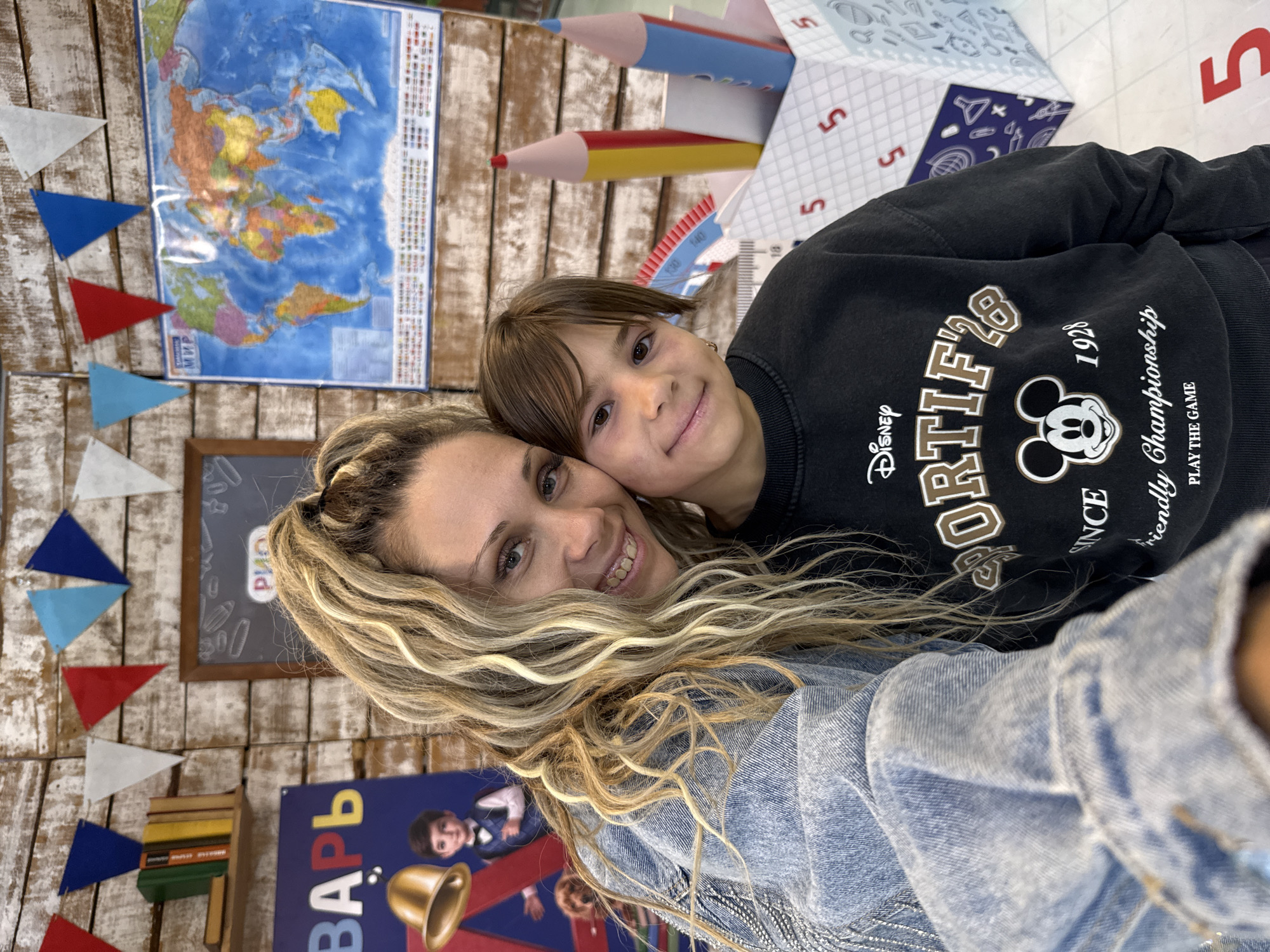 In Ignatovo, Russia, a woman with long blonde curly hair and a young child with dark hair are posing for a selfie. The woman is wearing a denim jacket, and the child is wearing a black sweatshirt with a Mickey Mouse graphic. Behind them, a wall is decorated with colorful triangular flags, a world map, and a chalkboard with writing. A sign featuring a bell and a child in a school uniform is also visible. The scene appears to be set up for a school-related event or celebration, possibly a "Day of Knowledge" or a back-to-school photo opportunity.