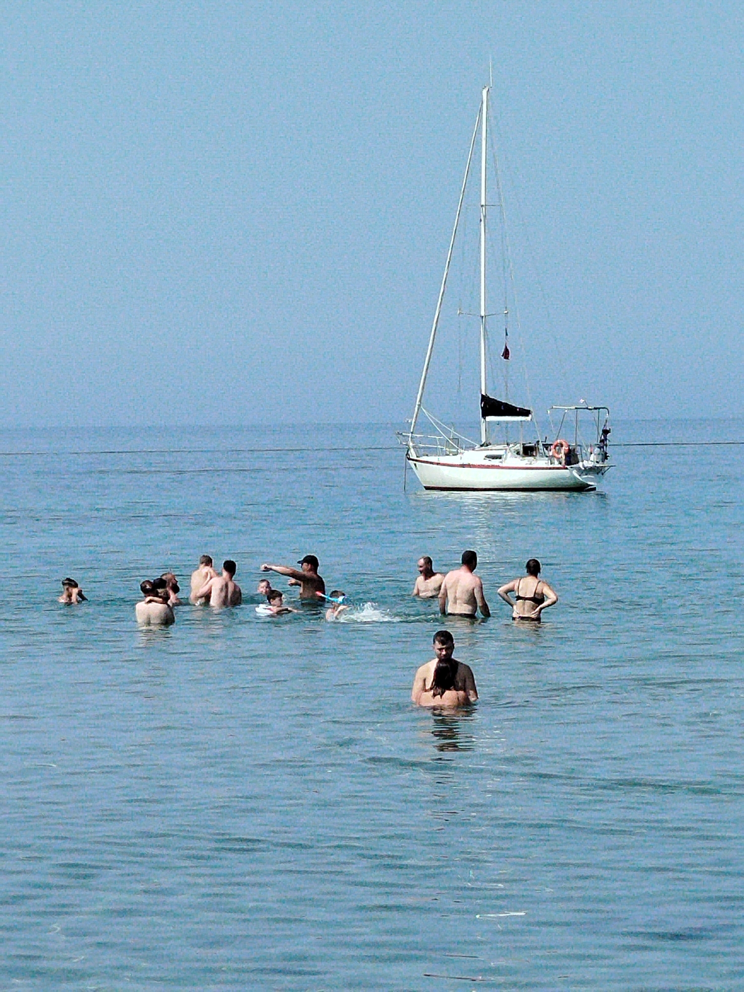 A group of people are swimming in the calm, blue waters of the sea near Sithonia Municipal Unit, Greece. A white sailboat with a black sail is anchored in the background. The sky is clear and blue. The people are scattered throughout the water, some are interacting with each other, while others are swimming independently. The water appears to be shallow enough for the swimmers to stand in. The scene depicts a peaceful day at the beach with a sailboat and swimmers enjoying the water.