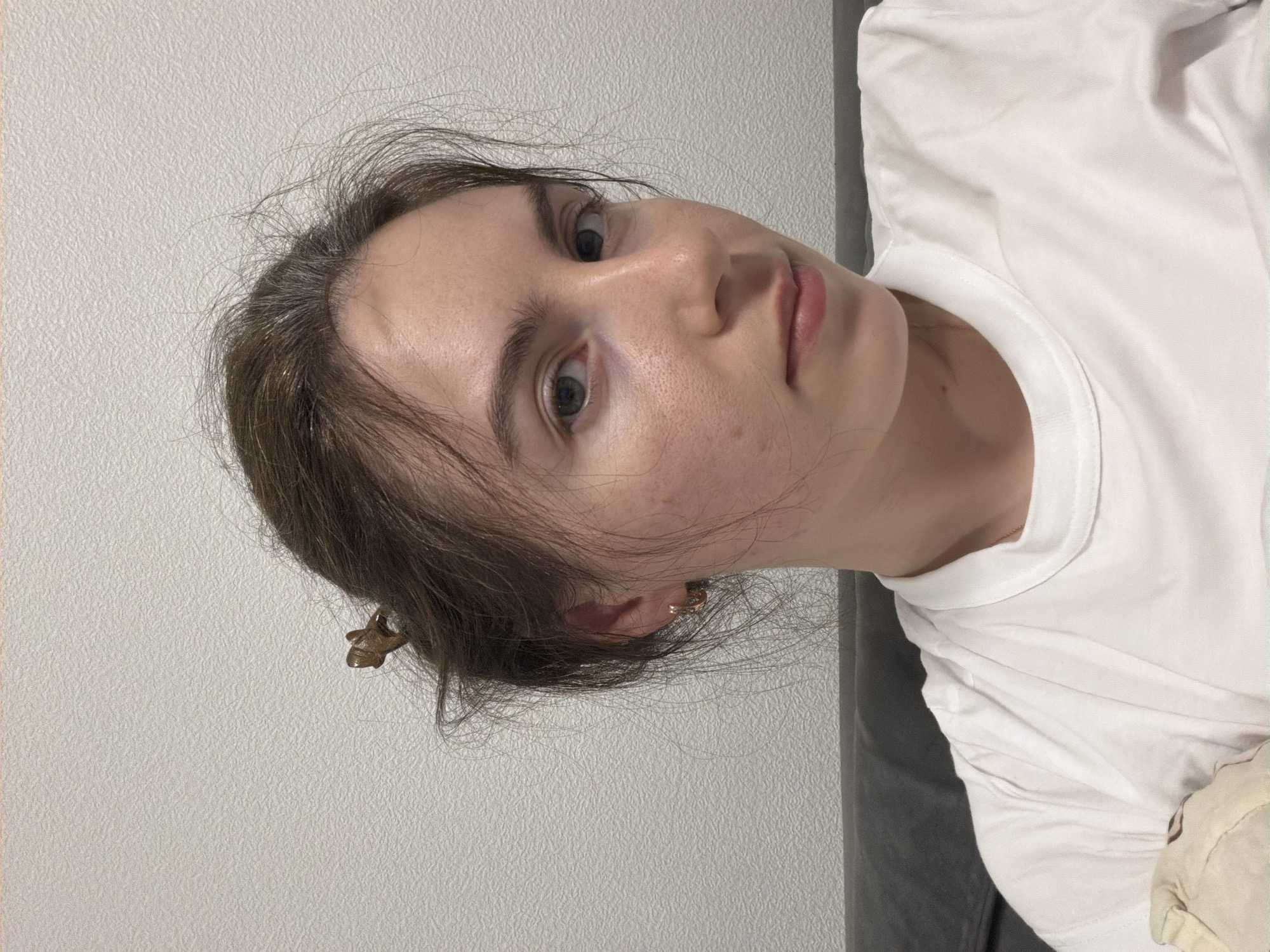 A young woman with fair skin and brown hair, partially pulled back, is facing the camera. She has blue eyes and her expression is neutral. She is wearing a white t-shirt. A gold earring is visible in her left ear. The background is a plain, light-colored wall. The image orientation suggests the photo was taken in portrait mode, possibly a selfie. No information indicates a location in Vsevolozhsk, Russia.
