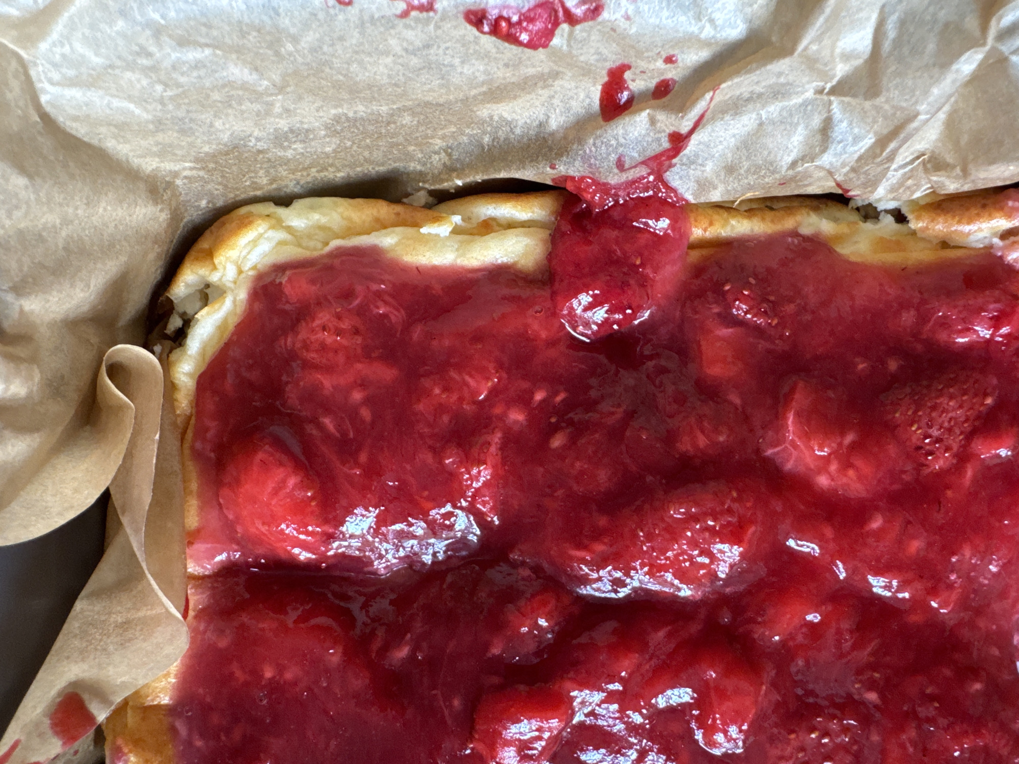 A close-up, overhead shot shows a baked good, likely a pie or cake, covered in a vibrant red fruit topping, possibly raspberries or strawberries, that has begun to seep over the edges. The baked good is set in a light brown parchment paper-lined dish. The texture of the fruit topping is thick and slightly gelatinous, with visible fruit pieces. The crust of the baked good is golden brown and appears to be slightly puffed. The parchment paper is crumpled in places and shows streaks of red fruit filling. The lighting is bright, creating highlights on the glossy surface of the fruit. The scene is devoid of human presence. The location of this image in Rzhev, Russia, is not visually inferable from the provided image.